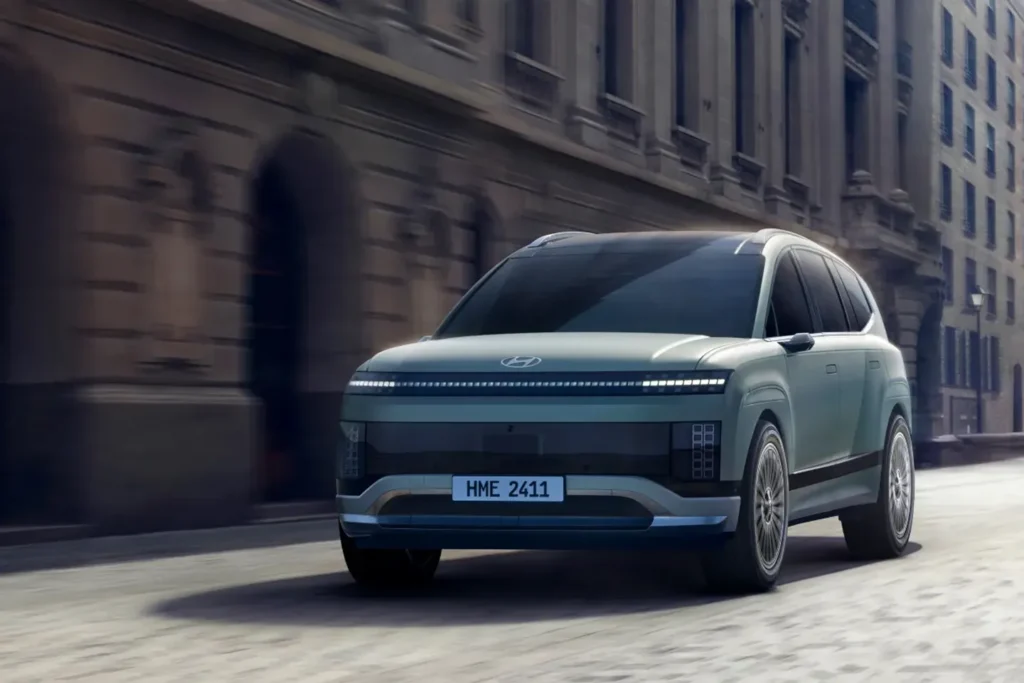 Hyundai Ioniq 9 electric SUV driving through a city street, featuring a minimalist front design with a full-width LED light bar.