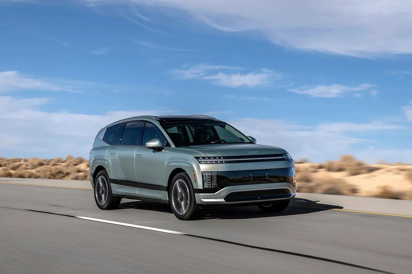 Hyundai Ioniq 9 electric SUV driving on an open road with dry scenery, showing its sleek aerodynamic design and LED front lighting.