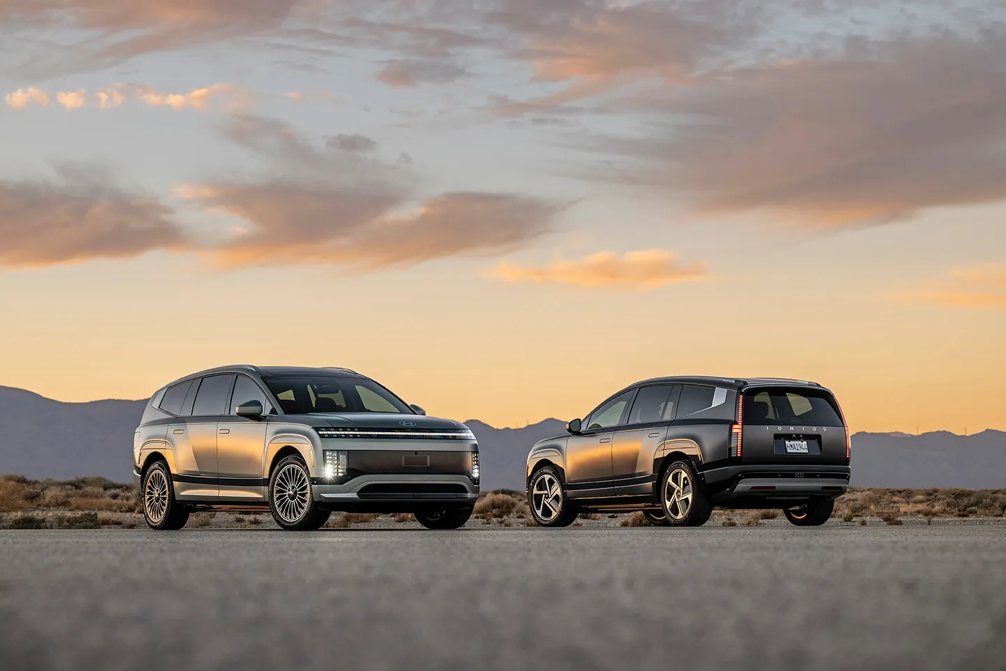 2026-hyundai-ioniq-9-sunset Front and rear three-quarter view of the Hyundai Ioniq 9 electric SUV parked in a dry landscape at sunset, highlighting its futuristic LED lighting and large three-row design.