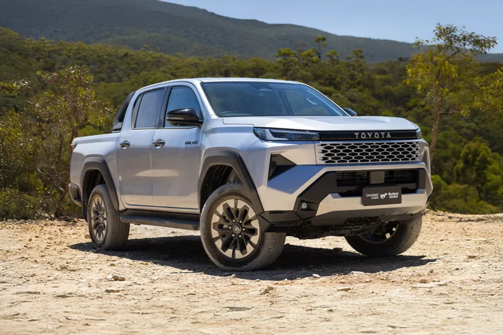 Silver Toyota Hilux pickup truck parked on a dirt clearing with forested hills in the background.