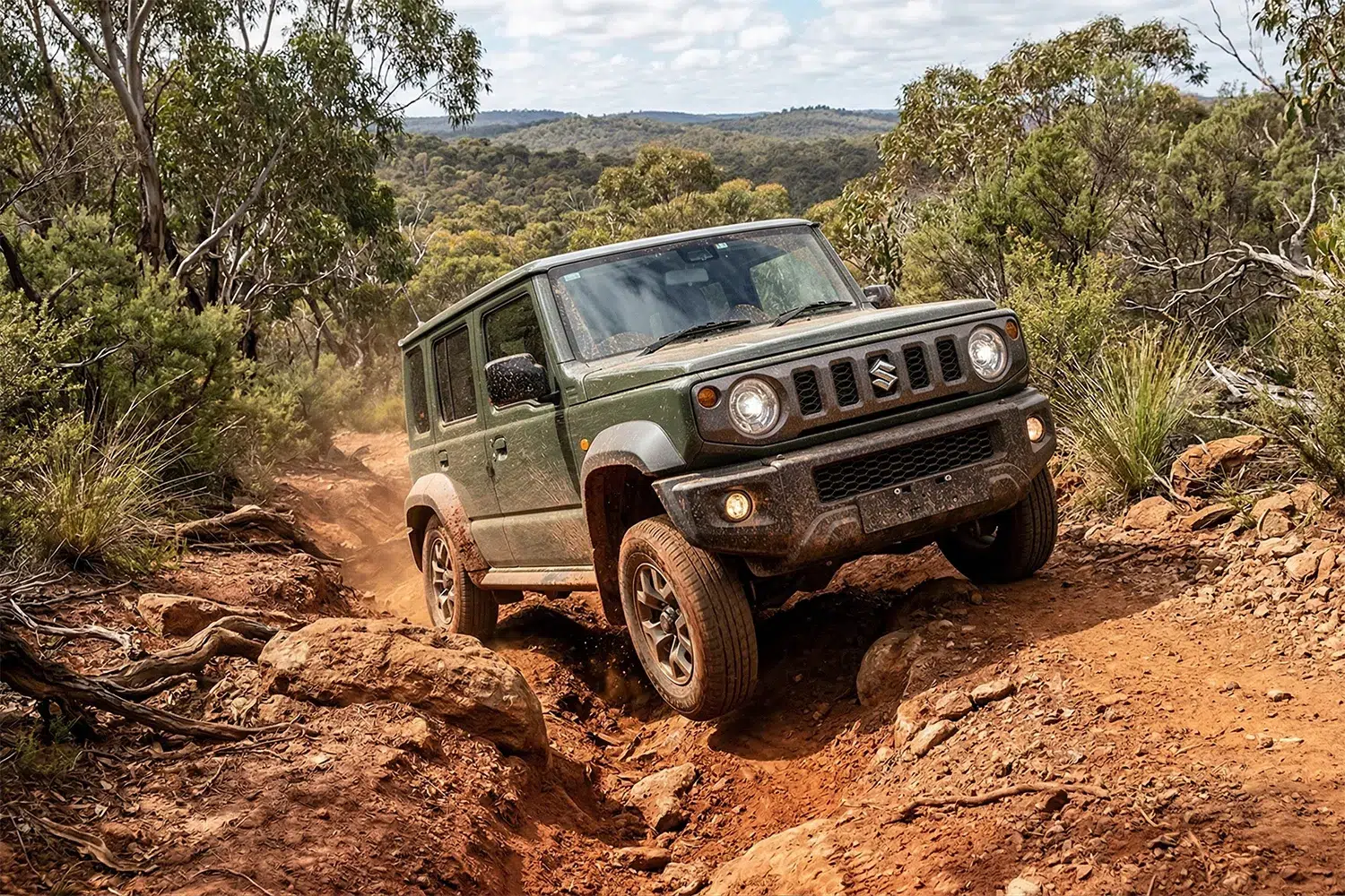 2026 Suzuki Jimny climbing a rocky red dirt trail off-road through Australian bush, front wheel articulating over a rock ledge