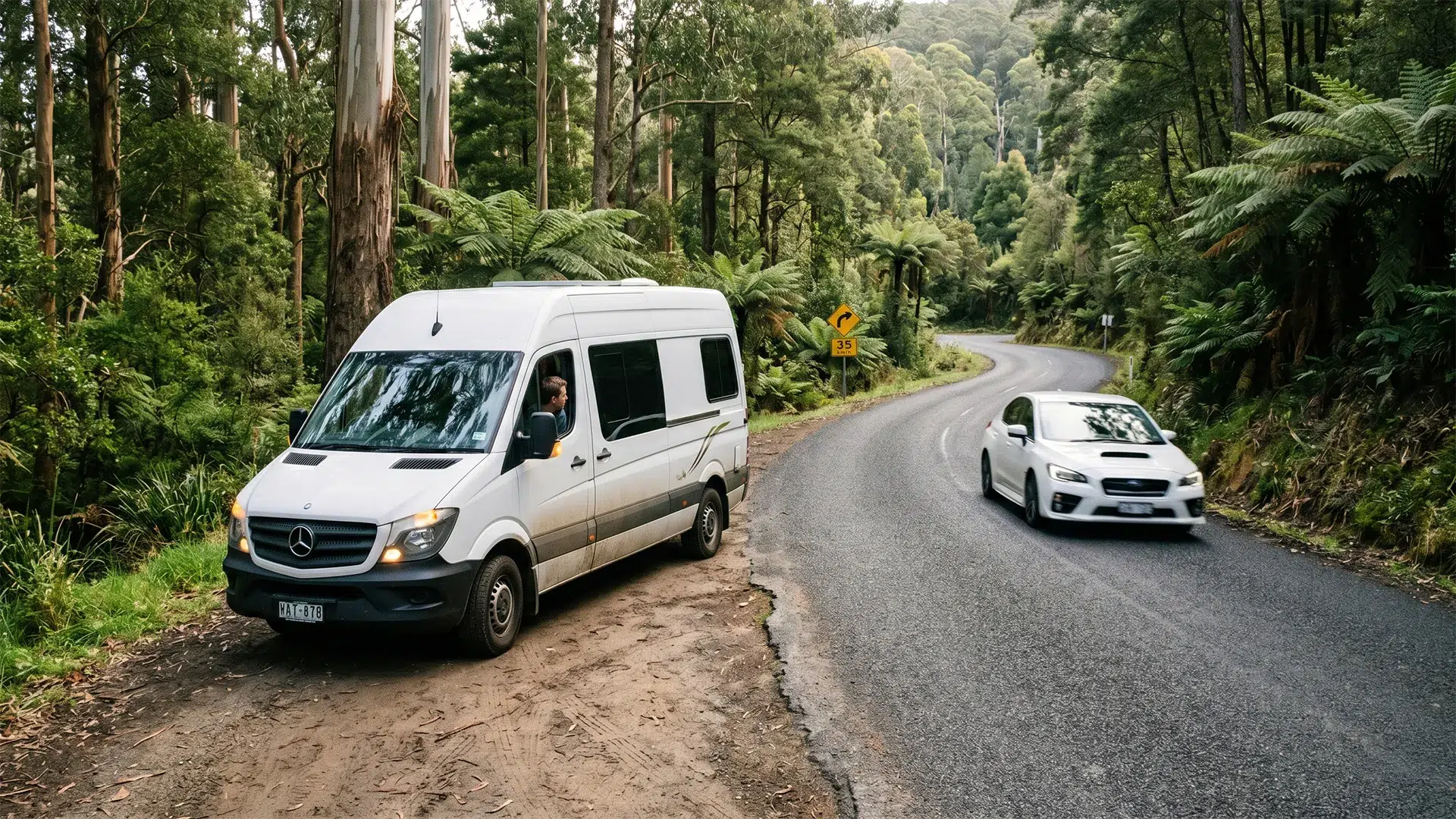 A white campervan pulled over onto a dirt shoulder to let a white Subaru WRX pass on a narrow winding Tasmanian forest road with a 35km/h advisory sign visible