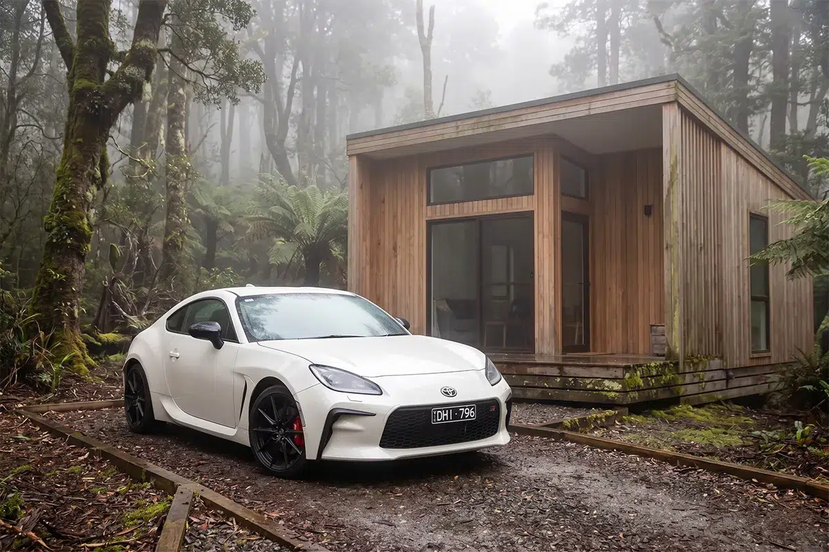 White Toyota GR86 parked on gravel outside a modern timber cabin surrounded by misty Tasmanian rainforest and tree ferns