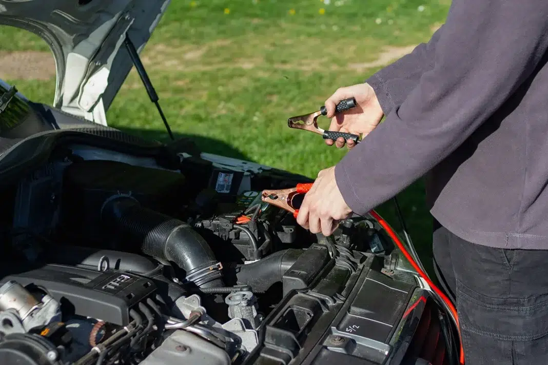 Person connecting red jumper cable clamps to a car battery in an open engine bay, jump starting a vehicle outdoors
