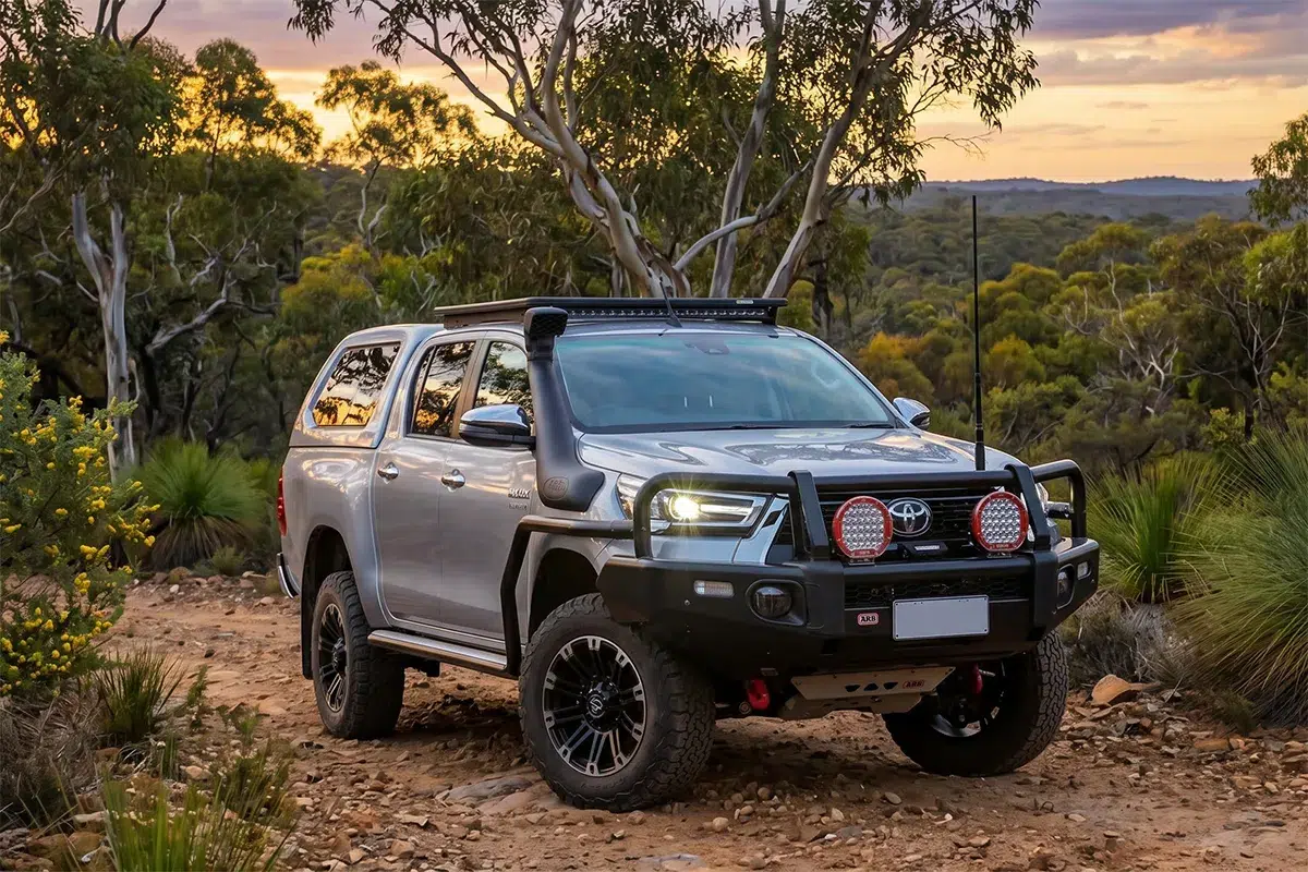 A silver Toyota HiLux fully outfitted with off-road accessories including a bull bar, prominent red driving lights, and a canopy, parked on a dirt trail in the Australian bush at dusk.