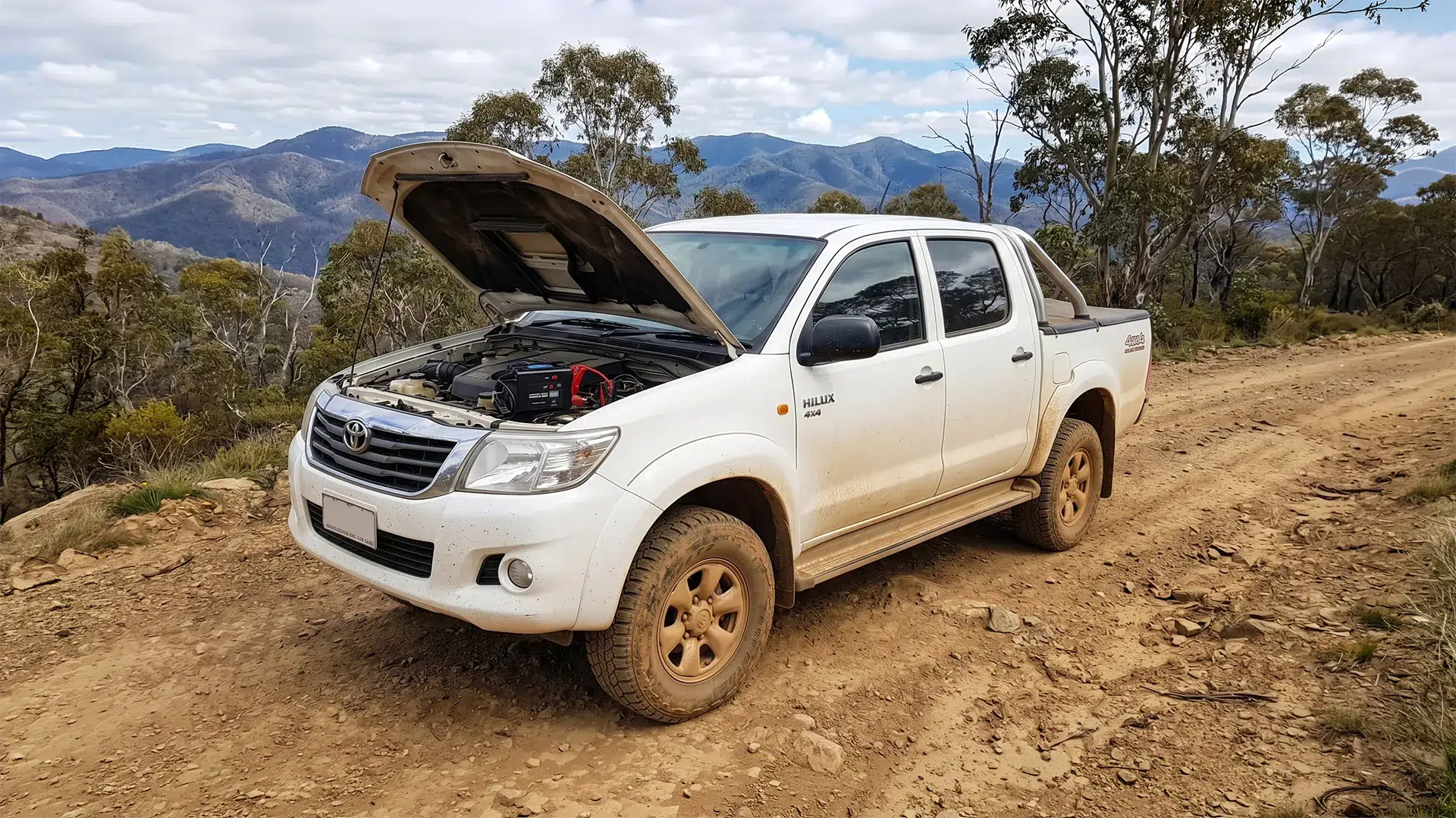 White Toyota HiLux 4x4 with bonnet open and jump starter connected, parked on a dusty dirt track in the Victorian High Country