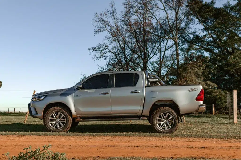 Side profile of a silver Toyota HiLux double-cab parked on a rural dirt track, equipped with all-terrain tyres and a sports bar.