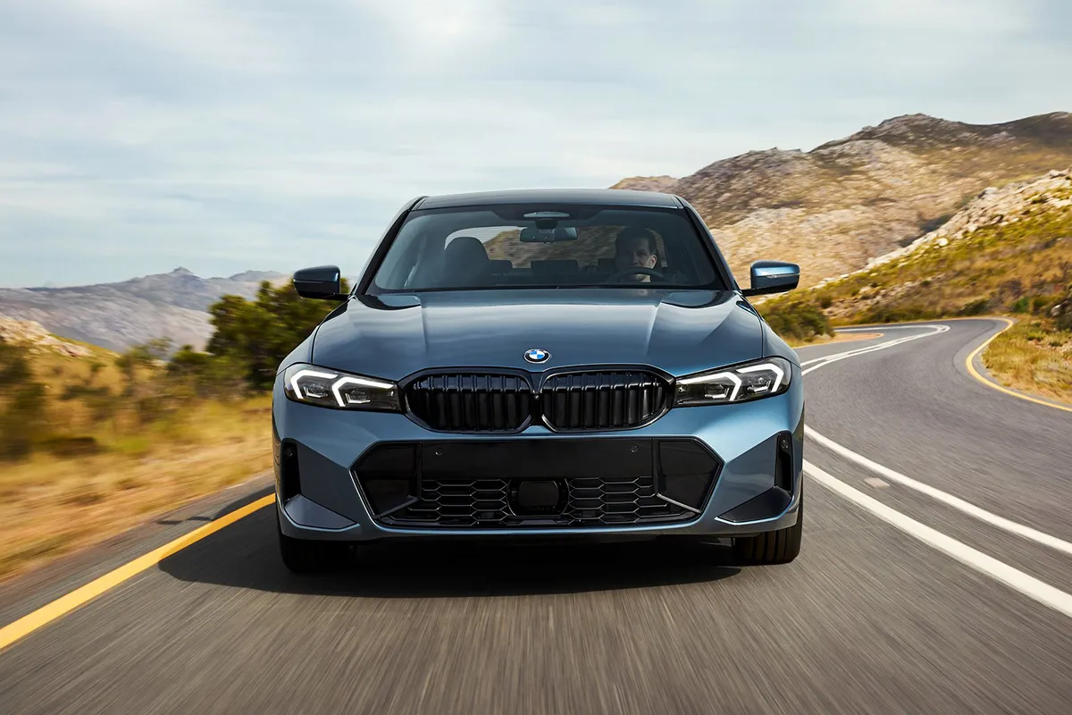 Blue 2026 BMW 330i M Sport driving on a winding mountain road showing the signature kidney grille and adaptive LED headlights