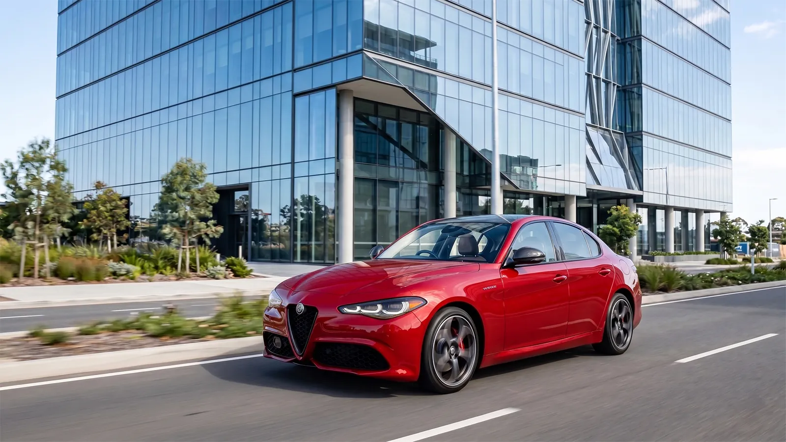 Red 2026 Alfa Romeo Giulia Veloce driving past a modern glass office building on an Australian suburban road
