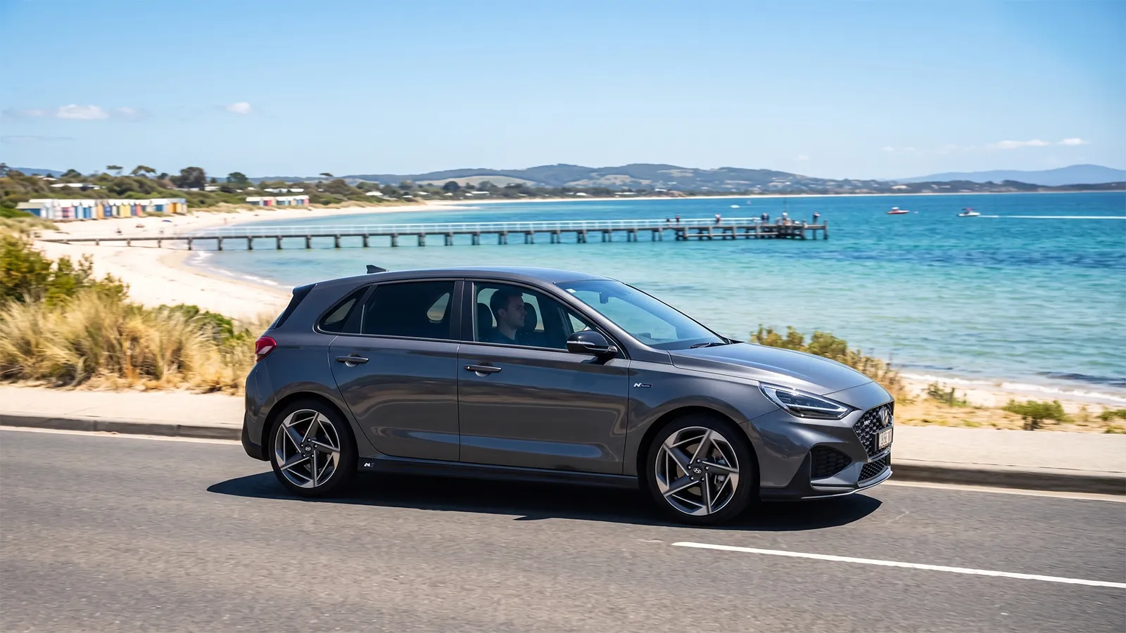 Grey 2026 Hyundai i30 N Line hatchback driving past a beach and pier on a sunny Australian coastal road