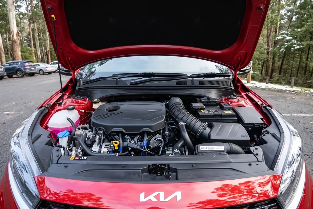 Engine bay of a red 2026 Kia Cerato GT showing the 1.6-litre turbocharged four-cylinder engine, photographed at an alpine car park