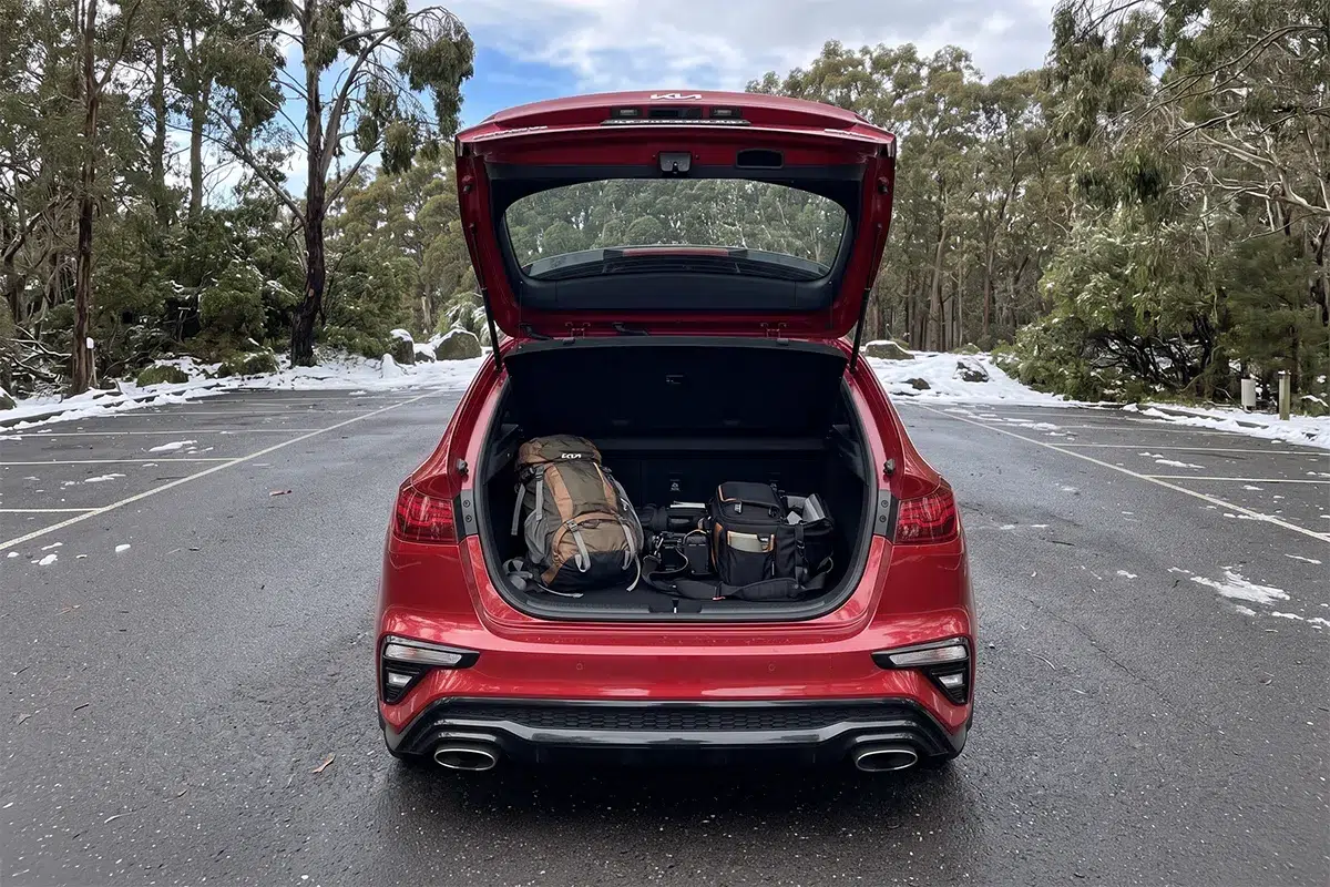 Open boot of a red 2026 Kia Cerato GT hatchback loaded with a hiking backpack and camera bags in a snowy alpine car park