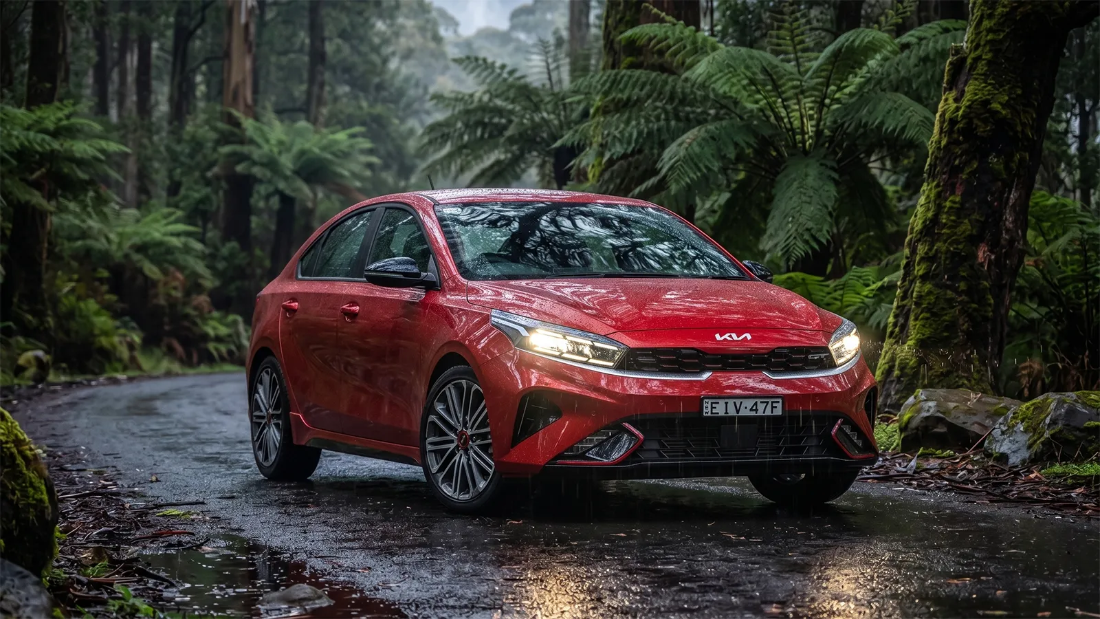 Red 2026 Kia Cerato GT hatchback parked on a wet rainforest road surrounded by tree ferns and moss-covered eucalypts