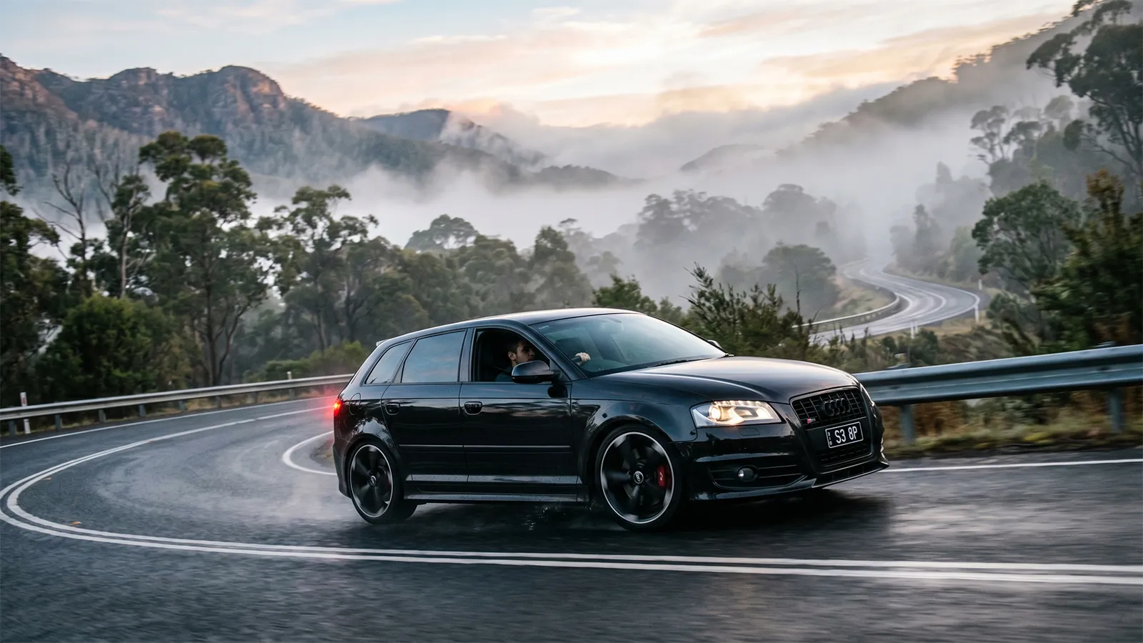 Black Audi S3 8P with "S3 8P" number plates cornering on a wet mountain road at dawn, mist rolling through forested valleys in the background.