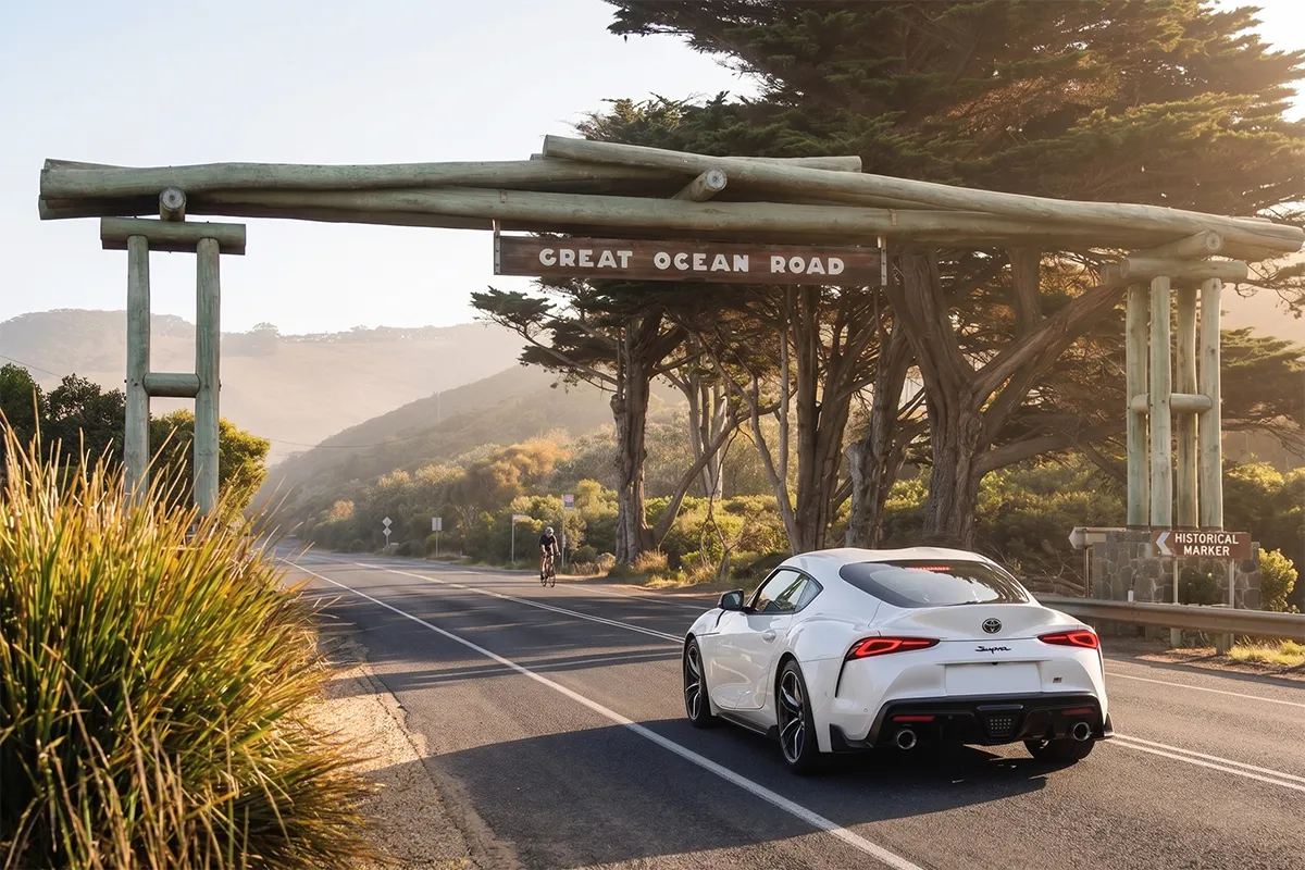 White Toyota GR Supra A90 driving beneath the iconic Great Ocean Road memorial archway at golden hour, with a cyclist passing in the distance.