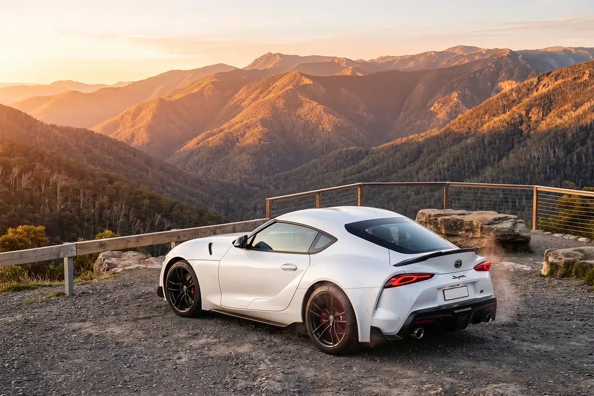 White Toyota GR Supra A90 parked at a mountain lookout at sunset, overlooking layered ranges of the Victorian Alps bathed in warm orange light.