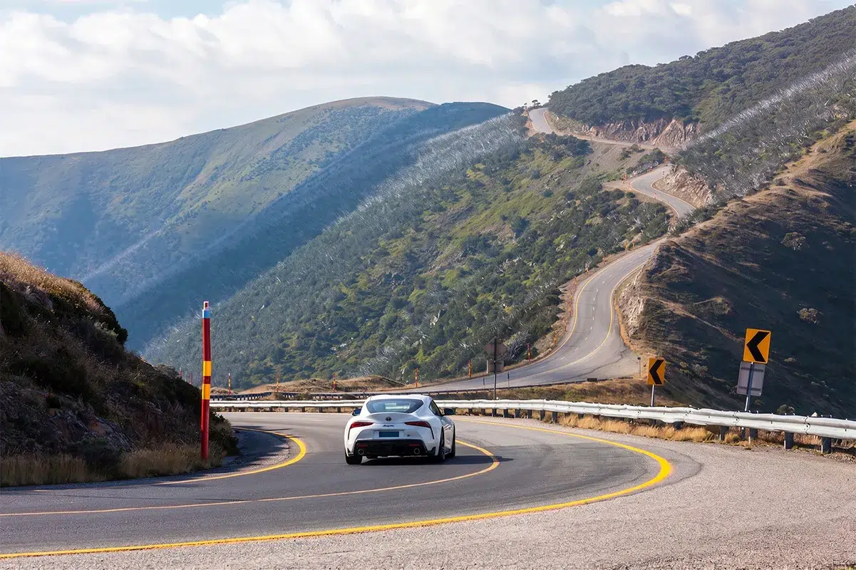 White Toyota GR Supra A90 cornering on the Great Alpine Road in Victoria, with the switchback road visible climbing the mountainside ahead.