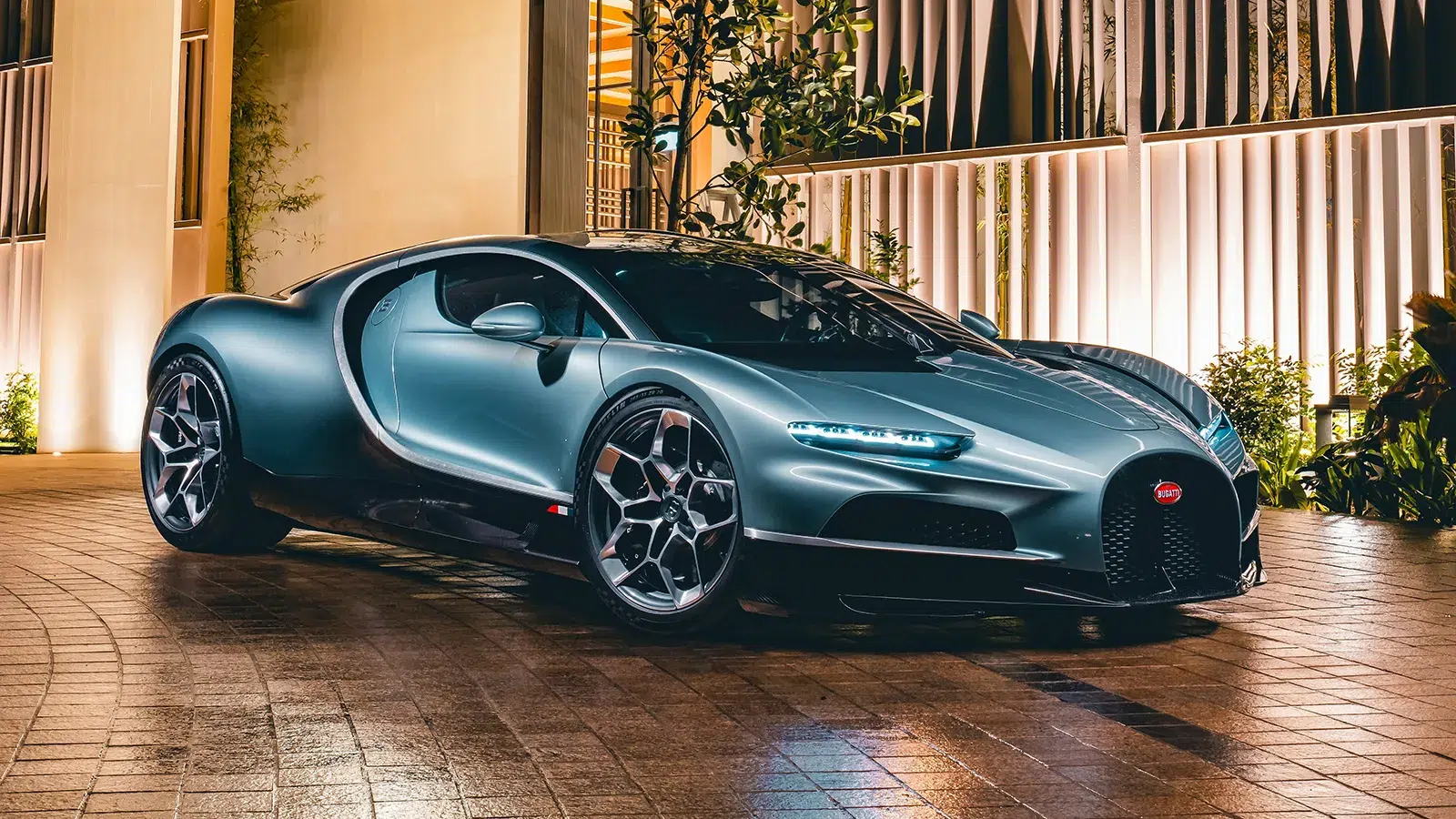 Bugatti Tourbillon in matte steel-blue parked in a luxury hotel forecourt at night, front three-quarter angle with warm ambient lighting and tropical foliage
