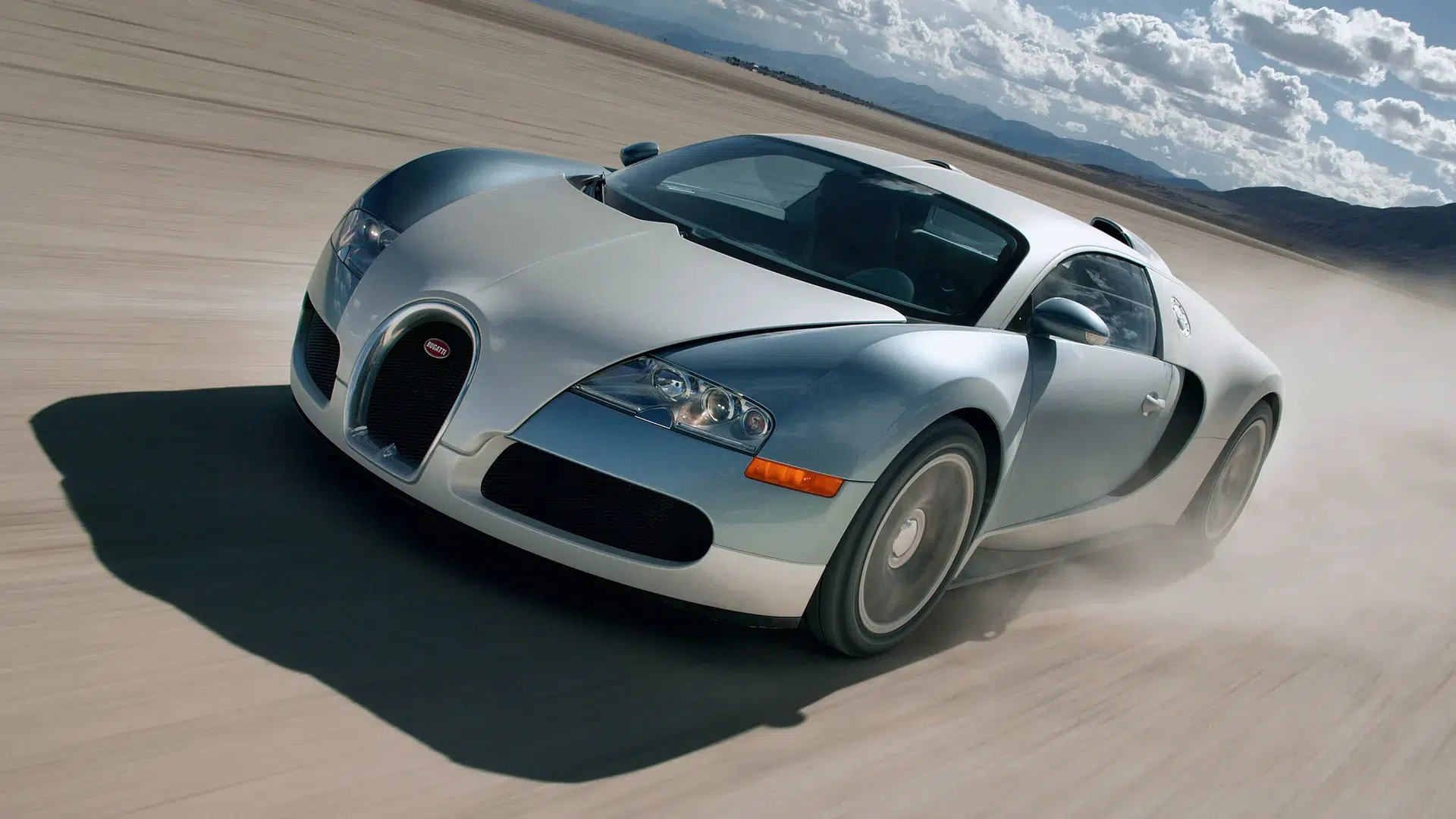 bugatti-veyron-16-4-lakebed-2005-front-three-quarter Bugatti Veyron 16.4 in silver-blue two-tone at speed on a dry lakebed, front three-quarter angle with motion blur and dramatic cloudy sky