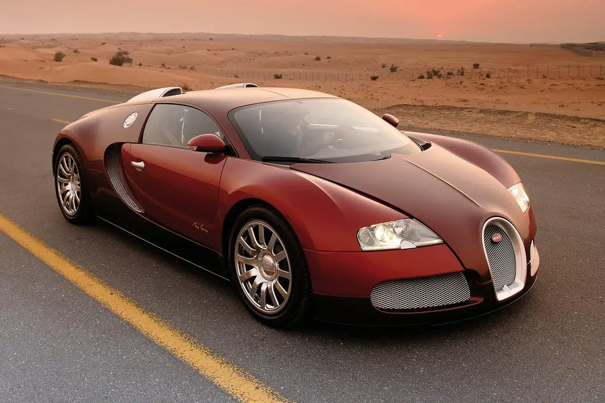 Bugatti Veyron in deep burgundy-red parked on a desert highway at sunset, front three-quarter with sand dunes and fading red sky in the background