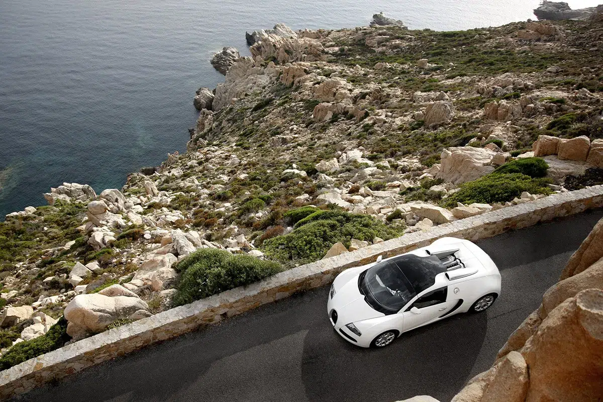 Bugatti Veyron Grand Sport in white shot from high above on a narrow clifftop coastal road, turquoise Mediterranean sea and rocky coastline below