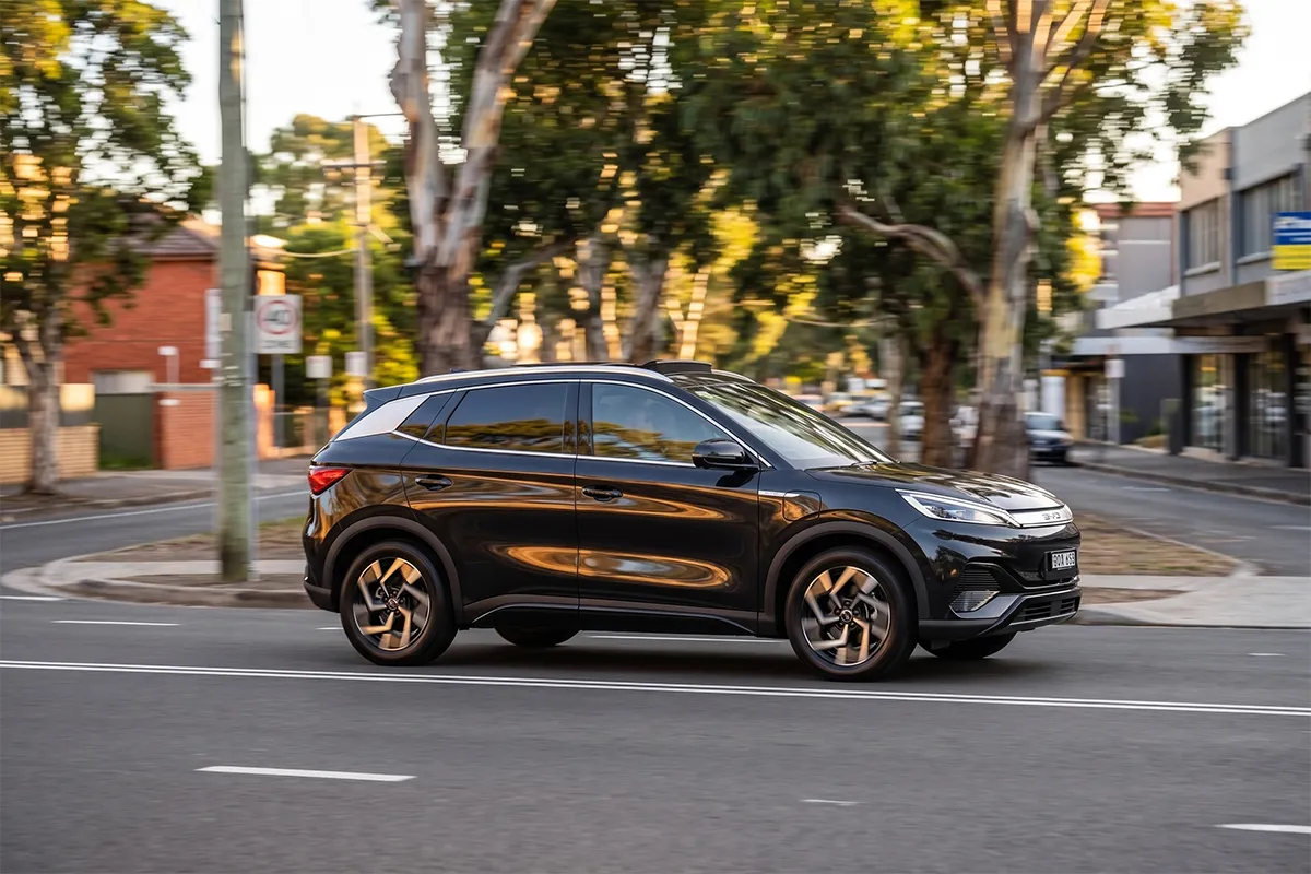 Black BYD Atto 3 driving along a tree-lined suburban street in side profile, showing its sloping roofline and alloy wheels