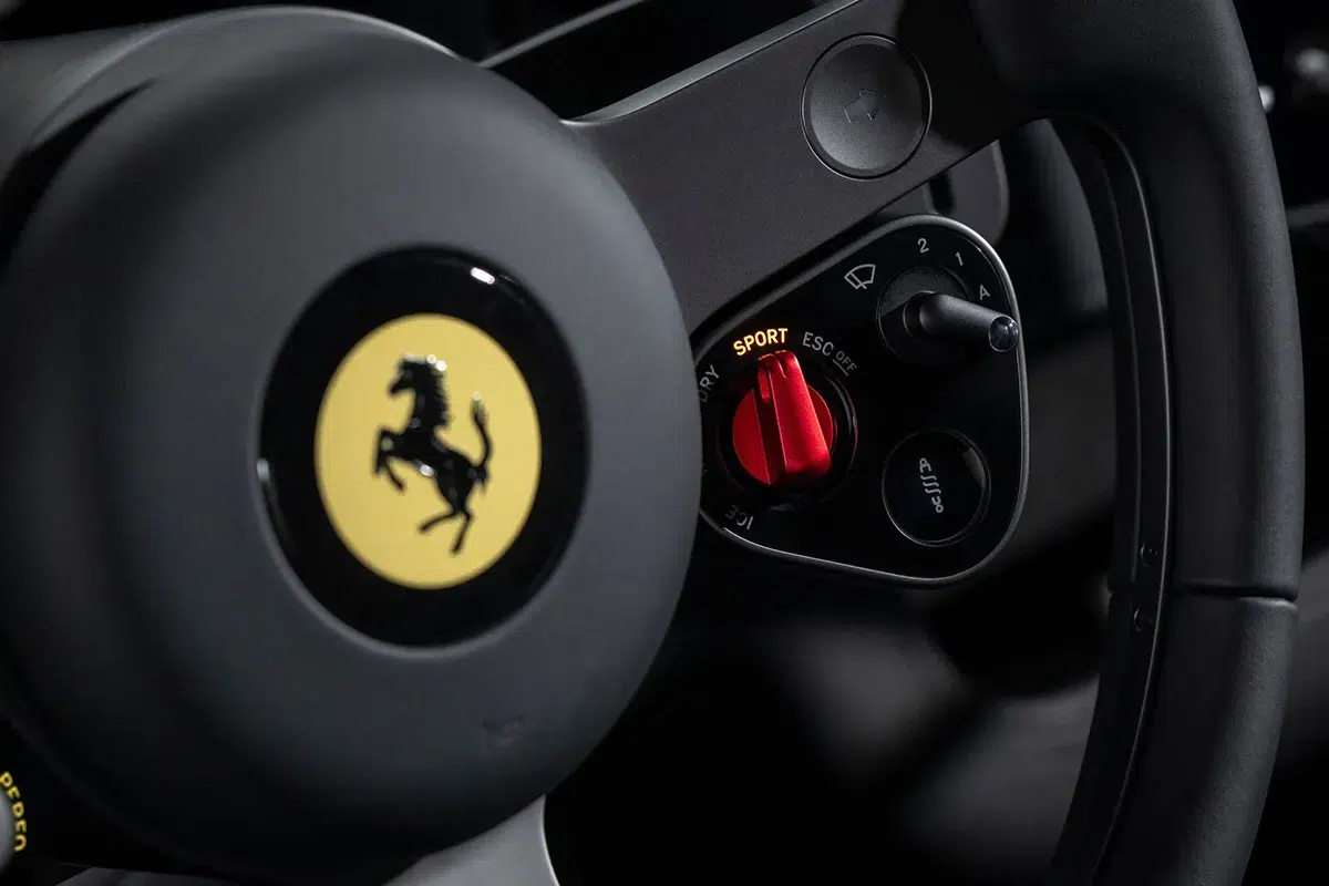 Ferrari Luce steering wheel close-up showing the red manettino dial set to Sport mode with ESC OFF and physical toggle switches