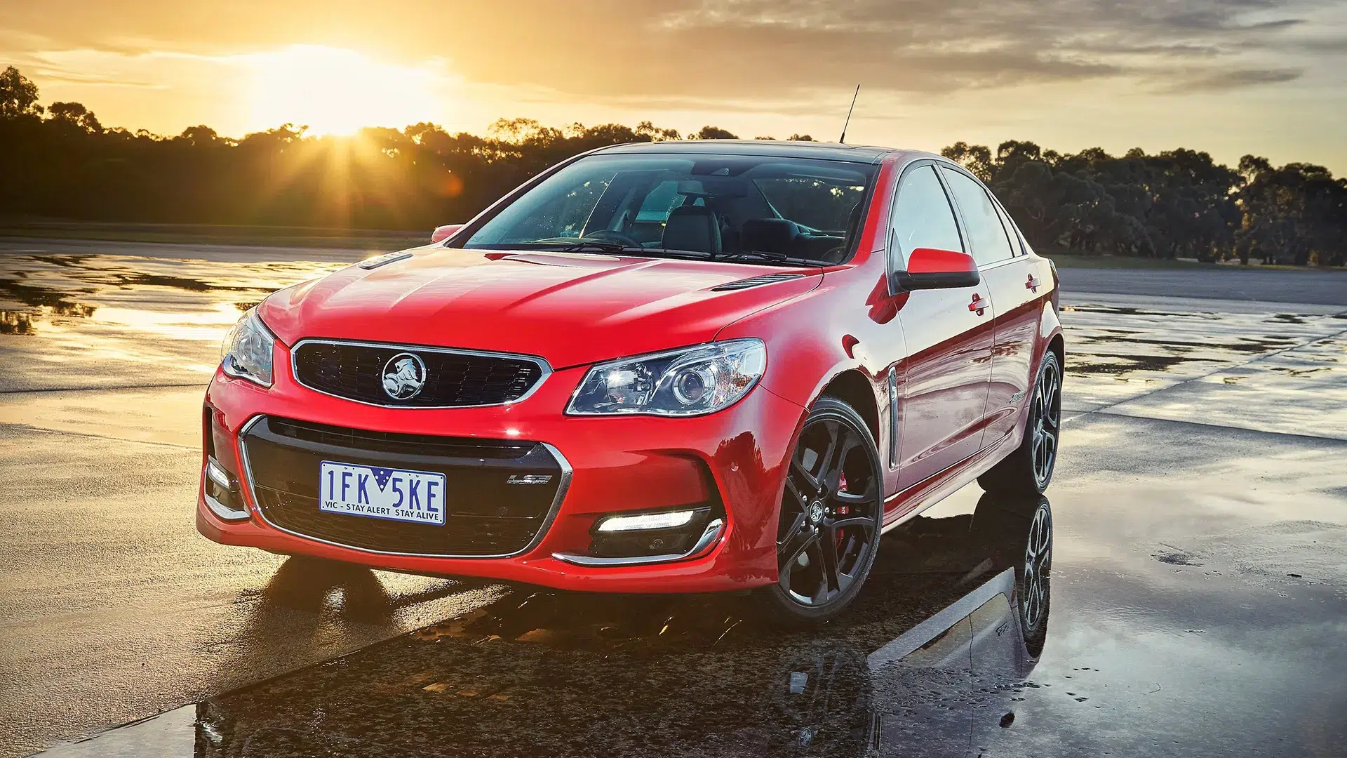 holden-vf-series-ii-commodore-ss-front-quarter-hero Red Holden VF Series II Commodore SS front three-quarter view at golden hour on wet tarmac with sunset sky