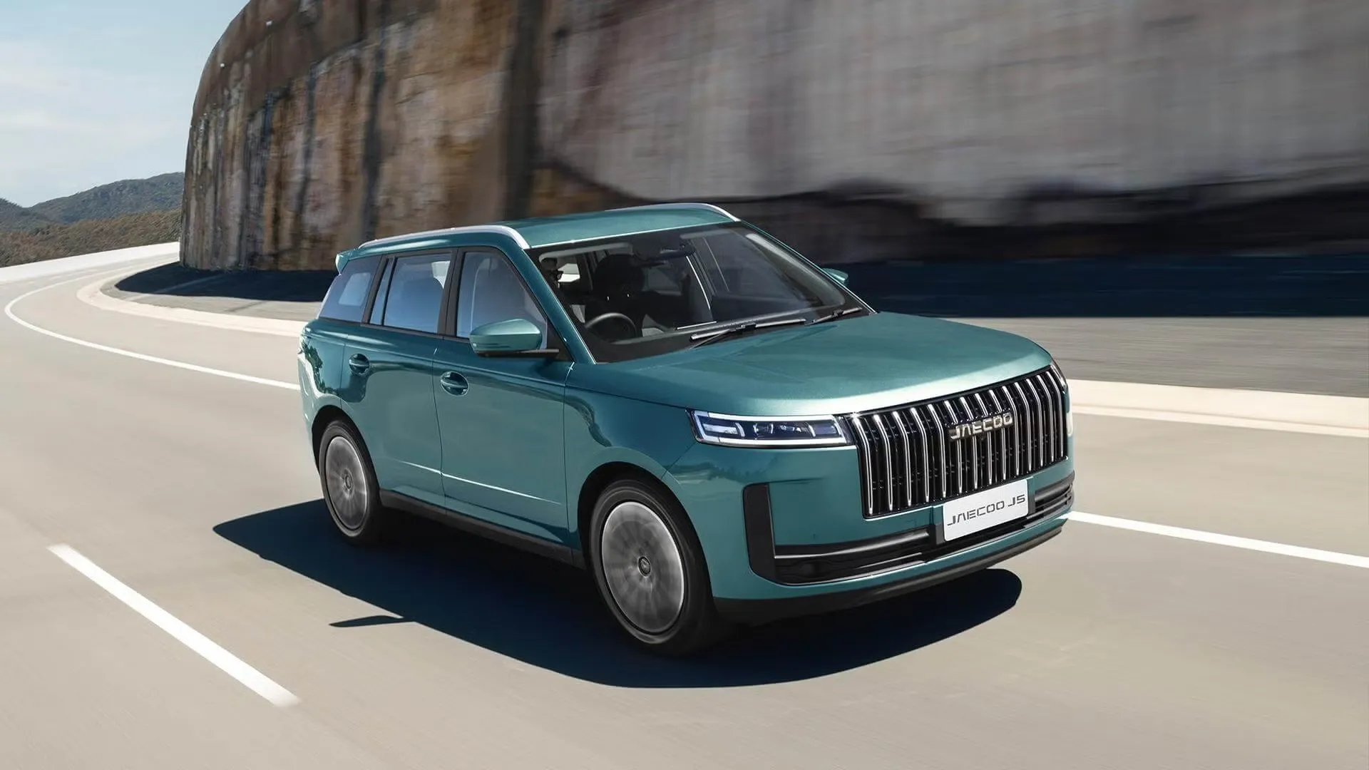 Teal Jaecoo J5 driving on a coastal mountain road, front three-quarter view in motion
