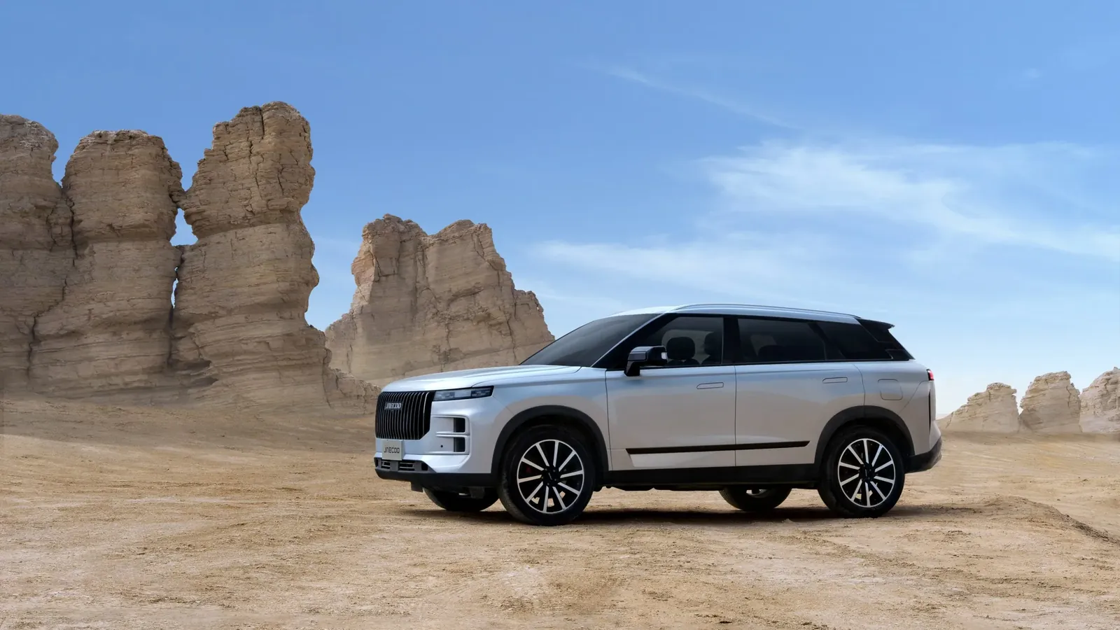 Silver Jaecoo J8 SUV parked on desert terrain beside weathered rock formations, side profile view