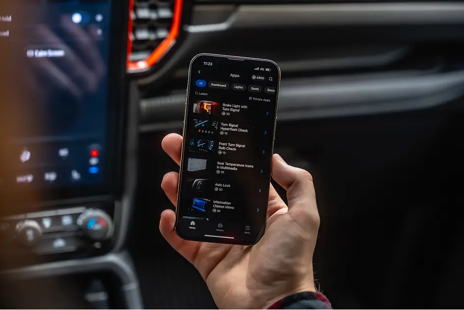 Hand holding a smartphone inside a car showing the OBDeleven app coding library with One-Click Apps for lights, seats and dashboard