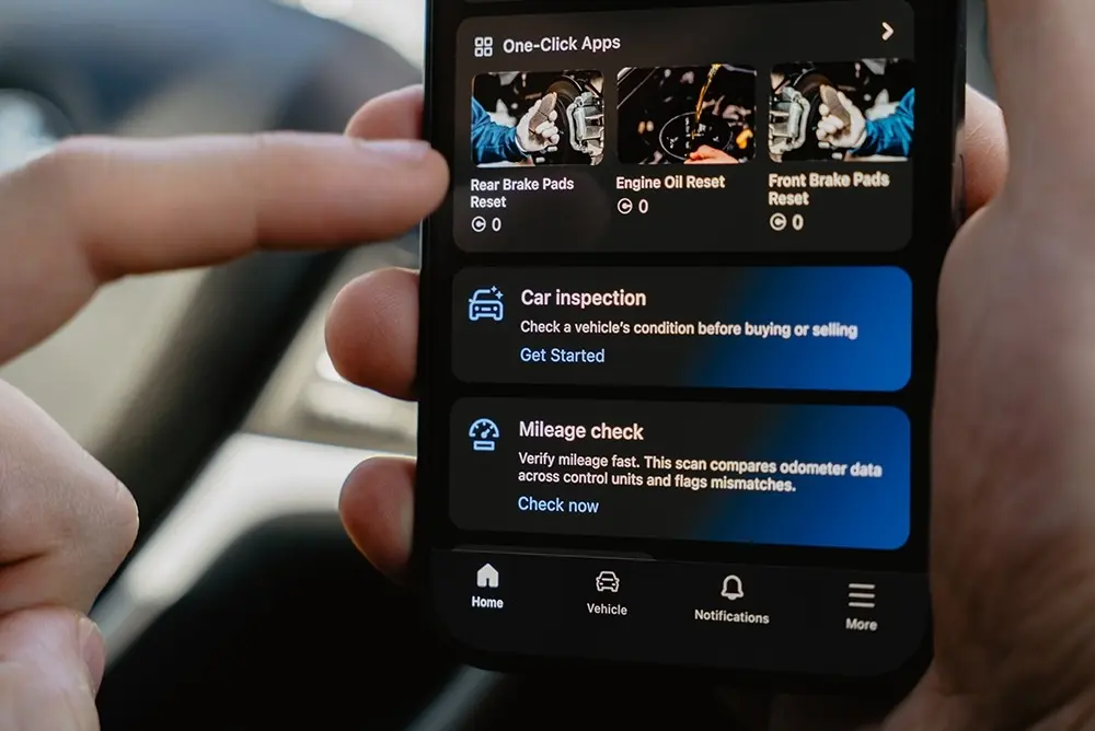 OBDeleven app displaying One-Click Apps including brake pad reset and engine oil reset on a smartphone held inside a car