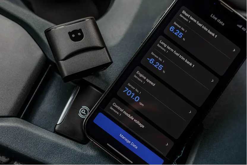 OBDeleven V3 adapter resting on a car centre console next to a smartphone showing live fuel trim and engine speed data
