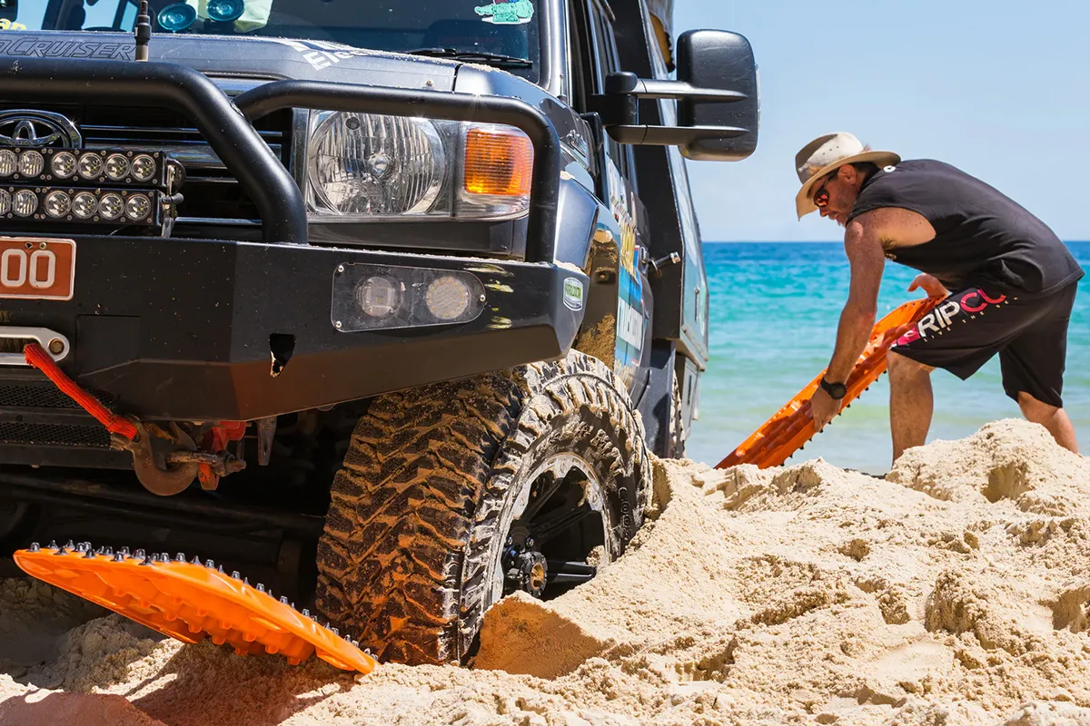 Person placing orange recovery boards under the front tyre of a bogged Land Cruiser on a beach
