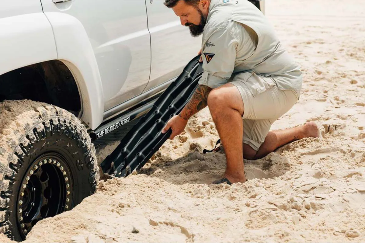 Person kneeling in sand to slide black recovery boards under the tyre of a bogged 4WD