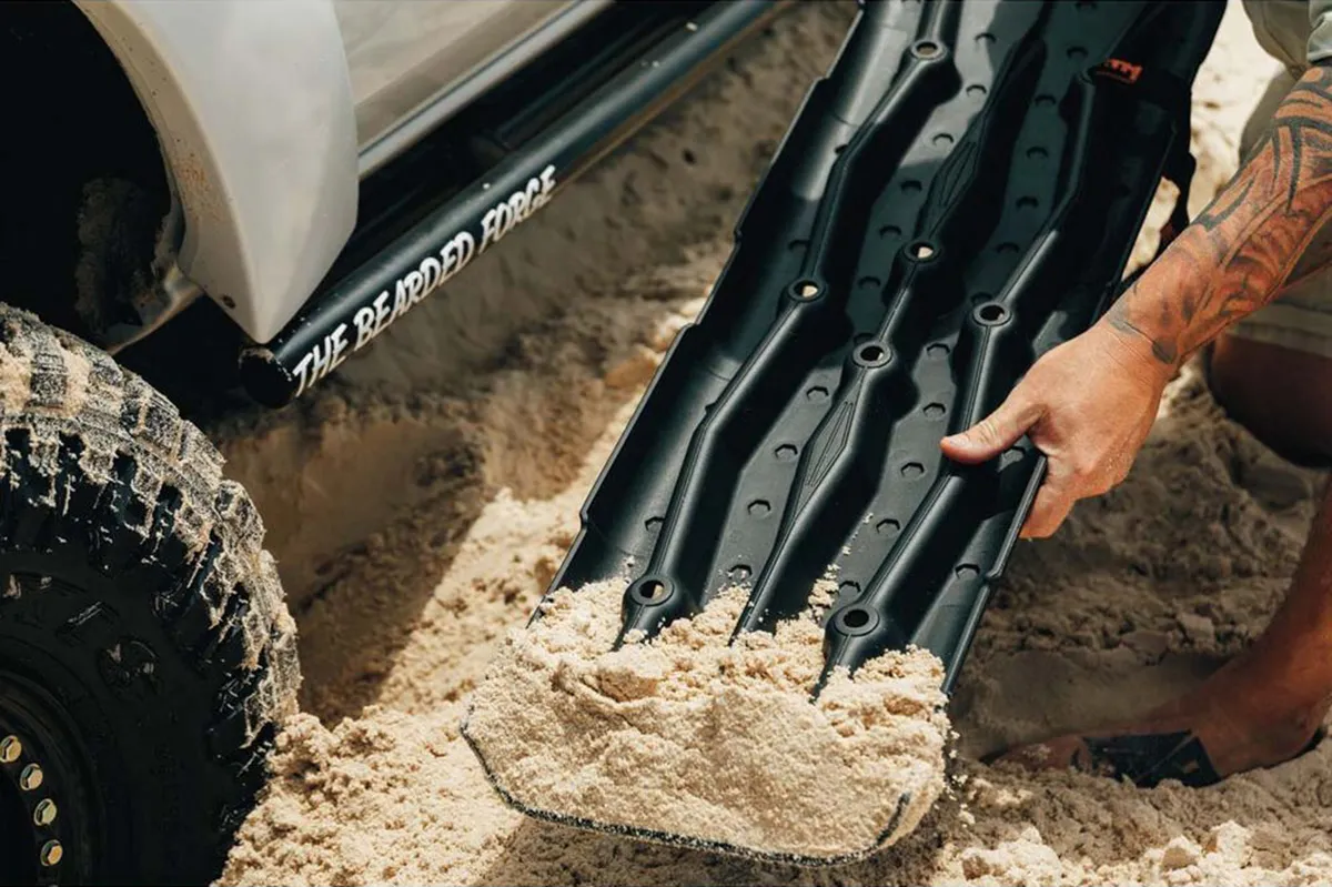 Person positioning black XTM recovery boards in sand beside a bogged 4WD tyre