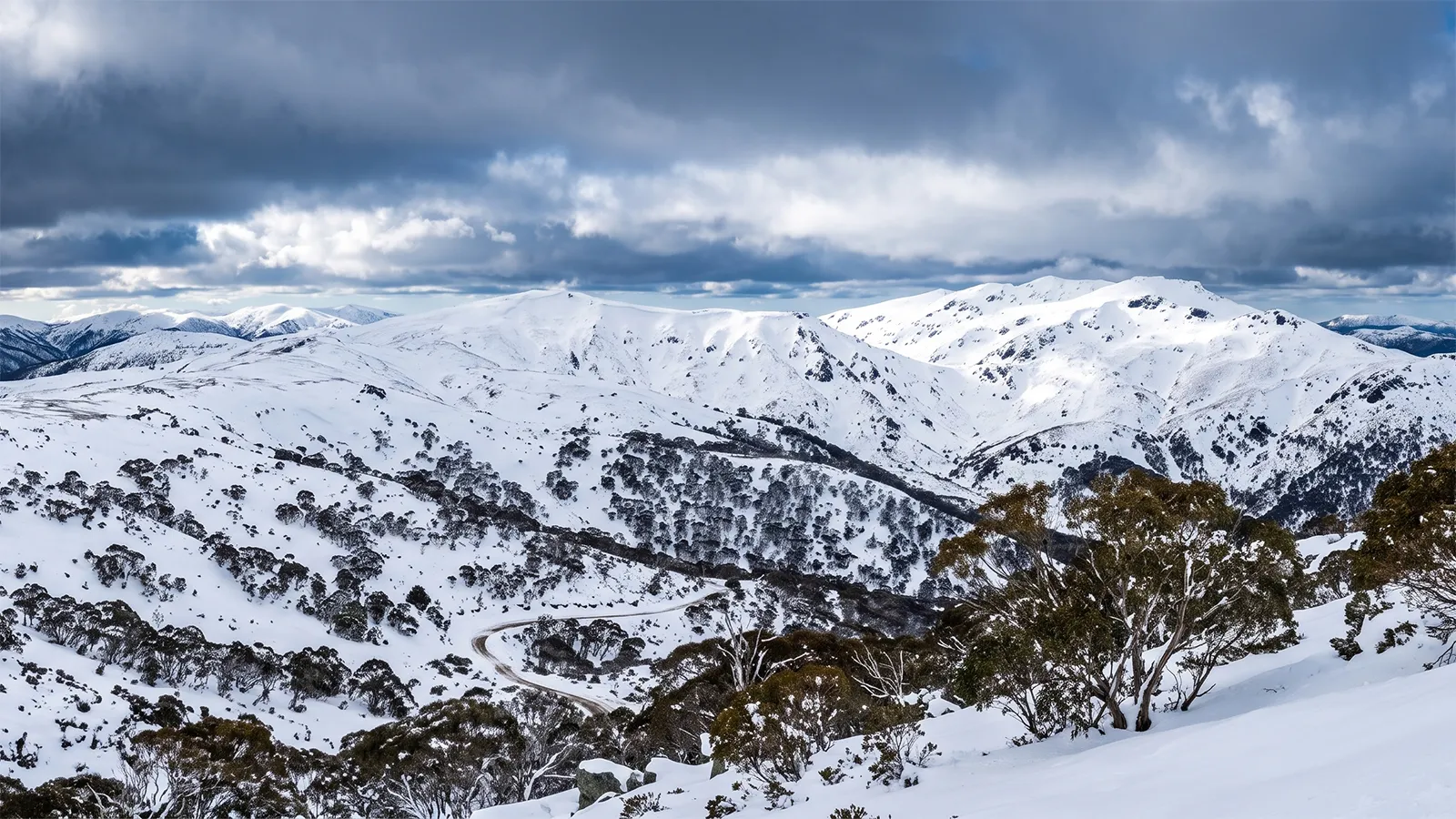 Panoramic winter view of snow-covered Snowy Mountains Main Range from high elevation with winding Alpine Way road visible in the valley below
