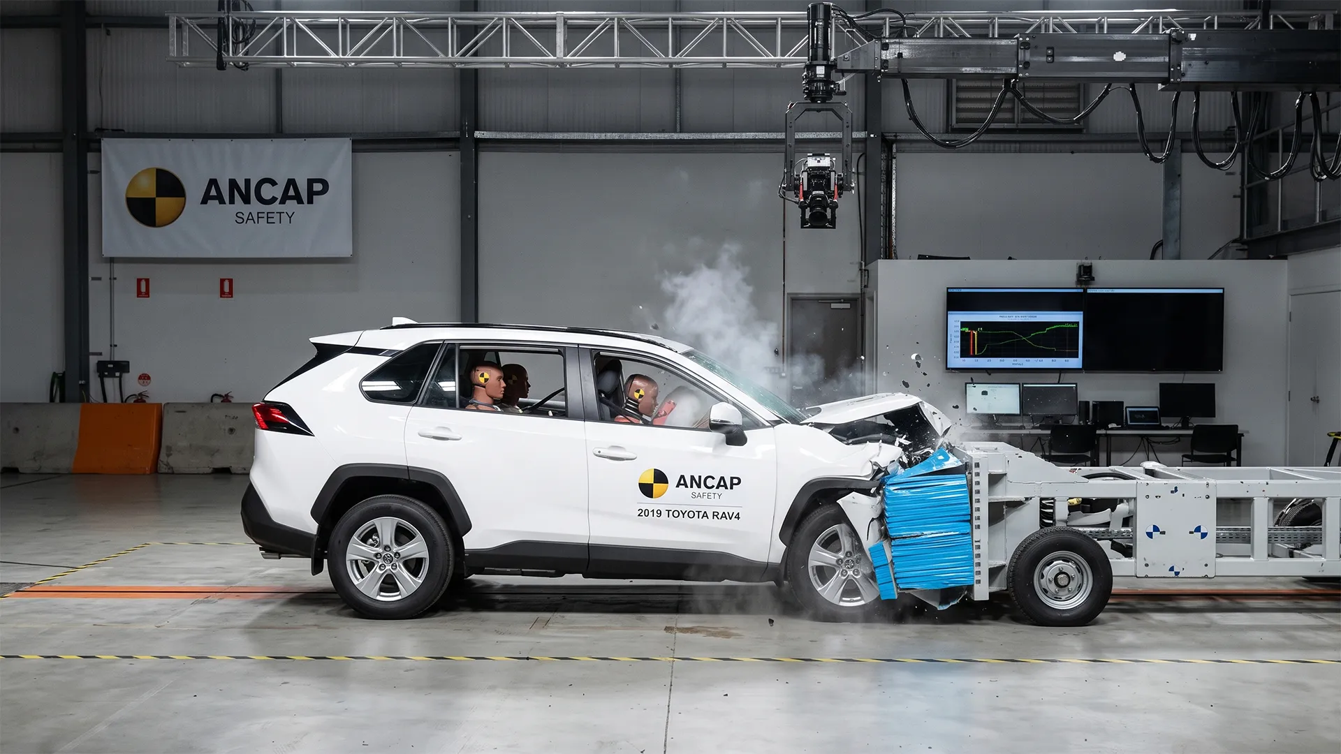 A front-impact crash test of a white Toyota RAV4 with test dummies, where the vehicle's front is damaged. Labeling on the car includes "ANCAP SAFETY" and the description "2019 TOYOTA RAV4".