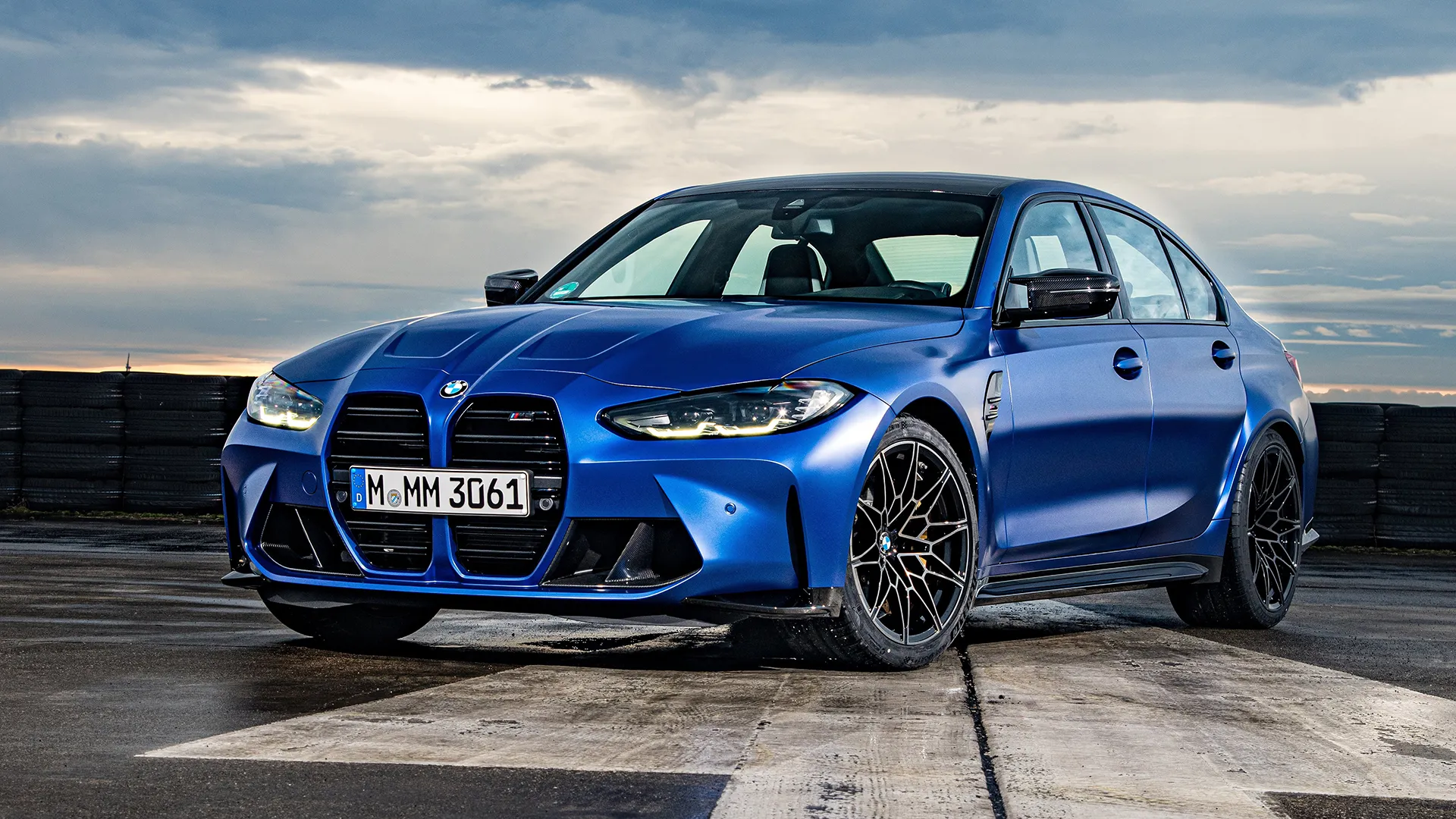 BMW M3 Competition in Portimao Blue on a wet track surface under dramatic overcast sky, front three-quarter view
