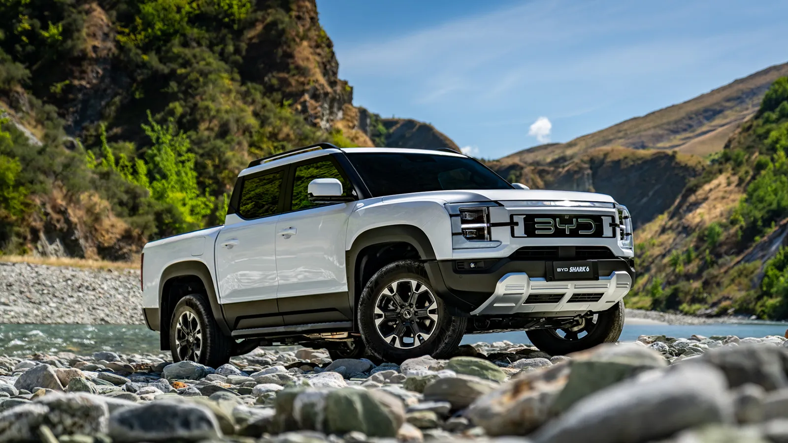 White BYD Shark 6 plug-in hybrid ute parked on a rocky riverbed below green mountains, front three-quarter view