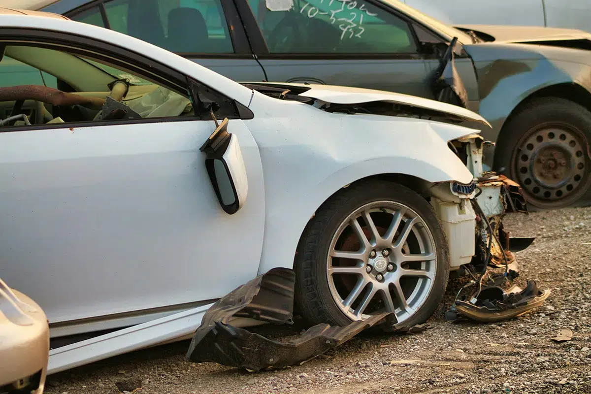 A white car with heavy front-end crash damage and a hanging bumper parked in a salvage yard at sunset