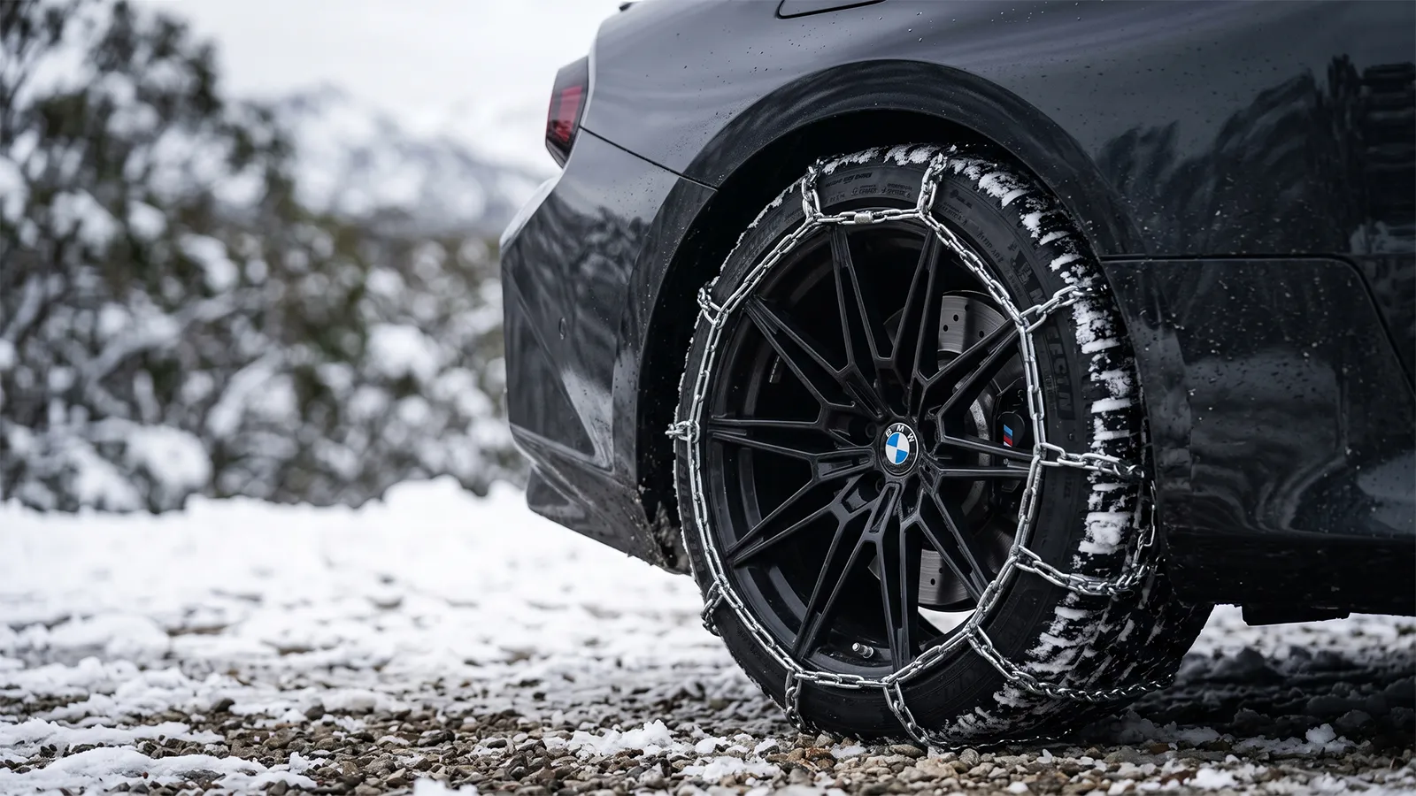 Snow chains fitted to the rear wheel of a BMW M2 G87 on a snowy alpine gravel surface, M-coloured brake caliper and BMW roundel visible through the spokes