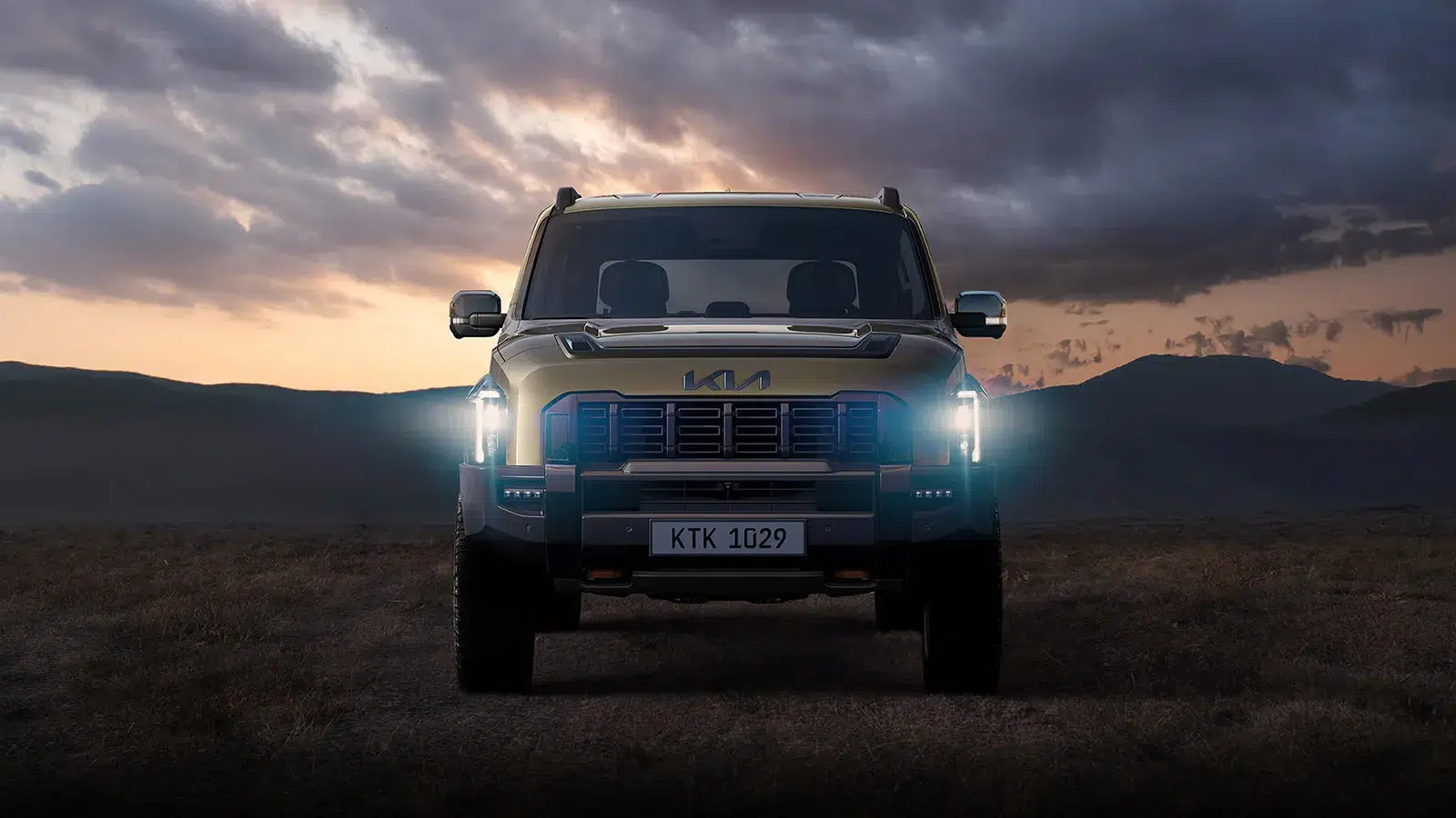 Kia Tasman X-Pro front-on with LED headlights glowing at dusk against mountains