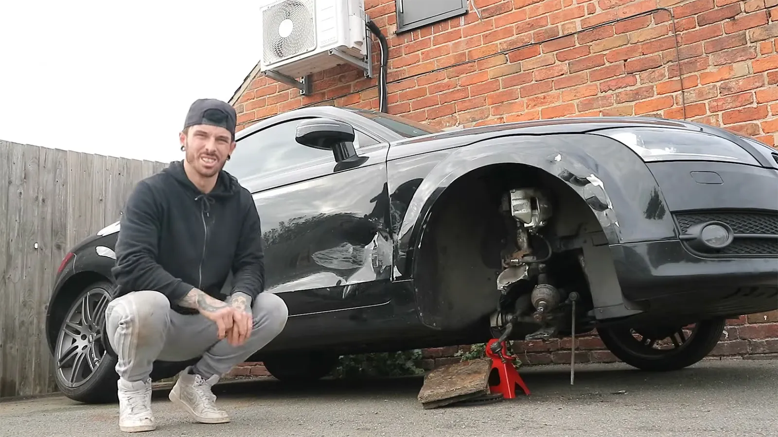 British YouTuber Mat Armstrong crouching beside a damaged black Audi TT with its front wheel removed during a driveway repair
