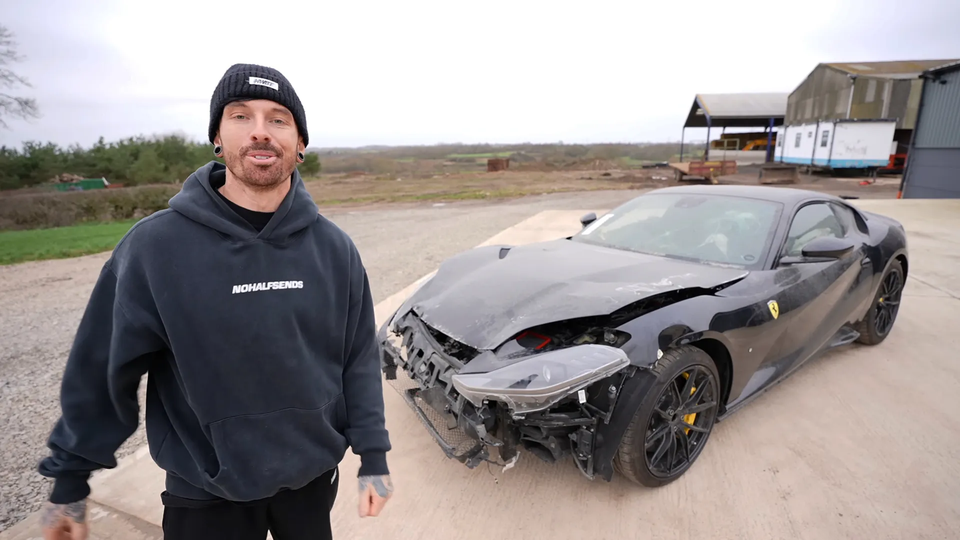 mat-armstrong-youtuber British YouTuber Mat Armstrong standing beside a crash-damaged black Ferrari with a smashed front end at a rural yard