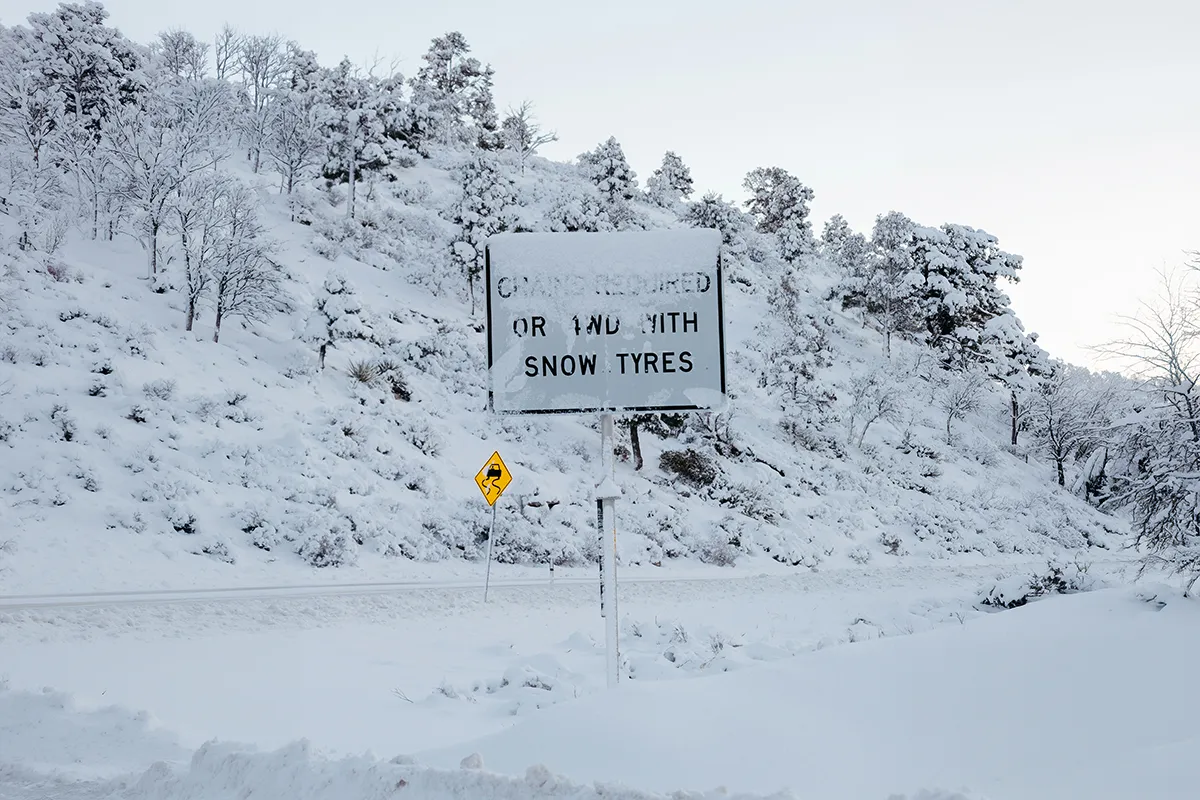 Snow chains required road sign partially buried in snow beside a mountain road in heavy winter conditions, slippery road warning sign