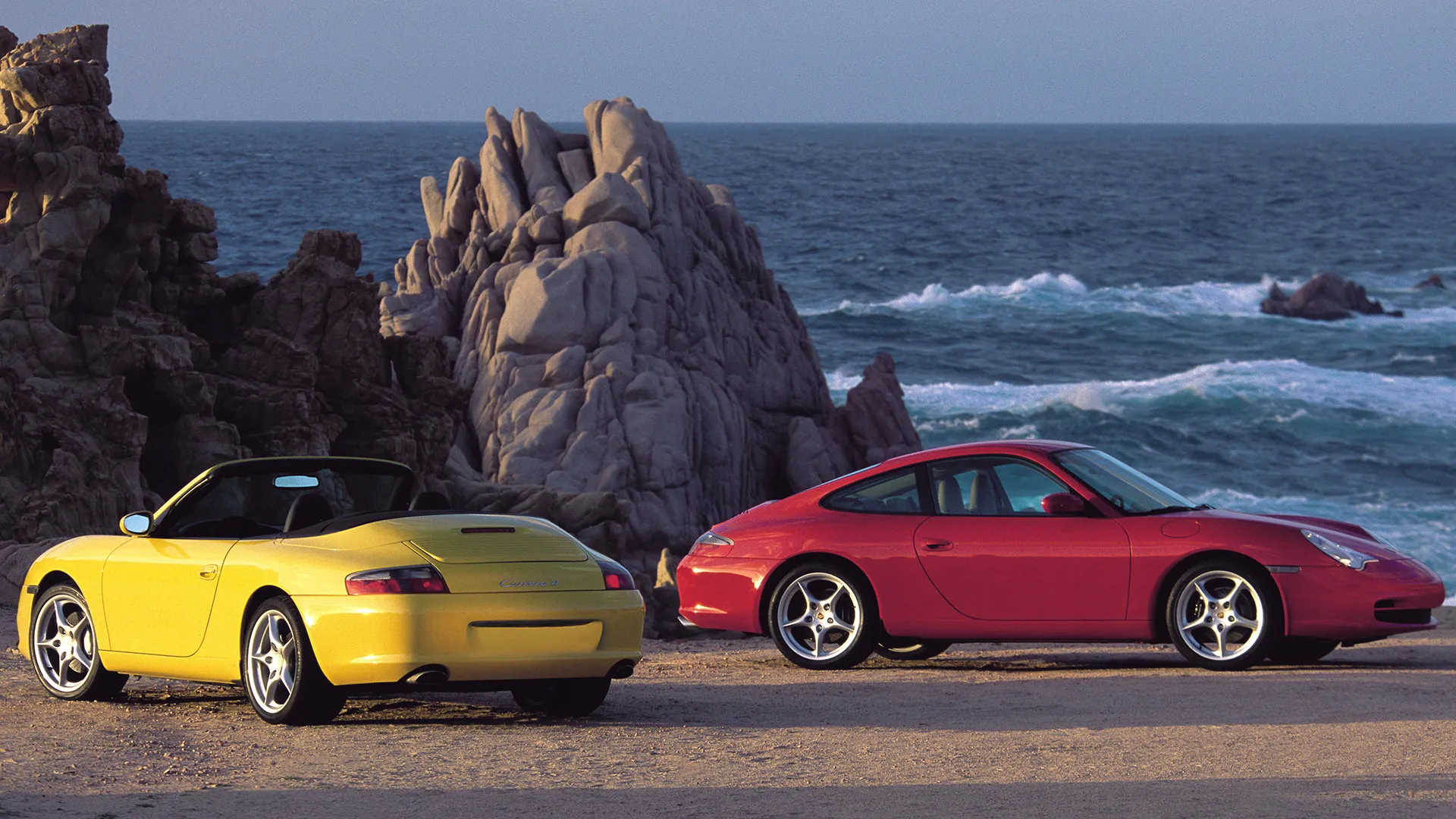 Yellow Porsche 911 996 Carrera 4 Cabriolet and red Carrera coupe parked on a rocky coastline