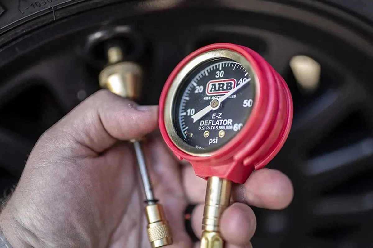 Hand holding a red and brass ARB E-Z Deflator with its analogue psi gauge beside a wheel