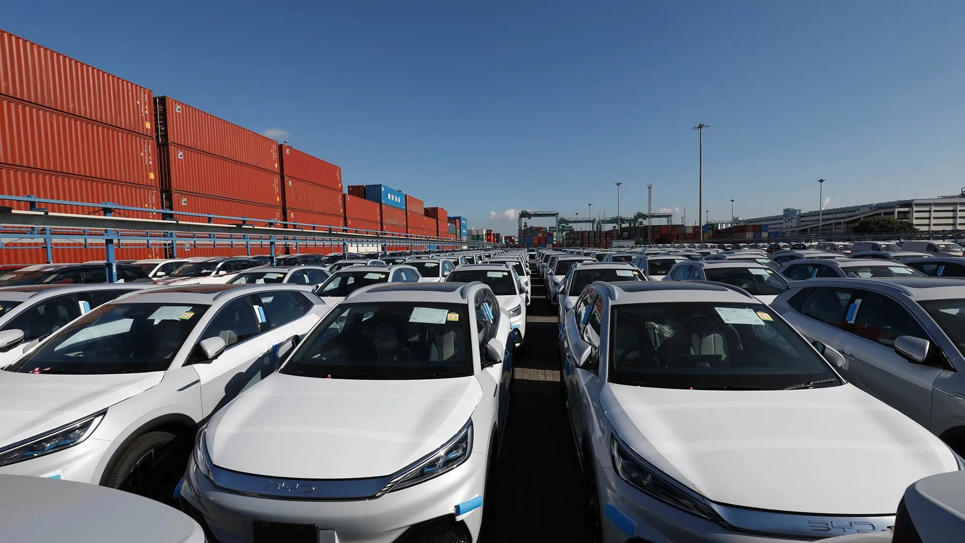 byd-ev-car-yard-aus Rows of new white electric SUVs parked at a shipping port with stacked containers behind them
