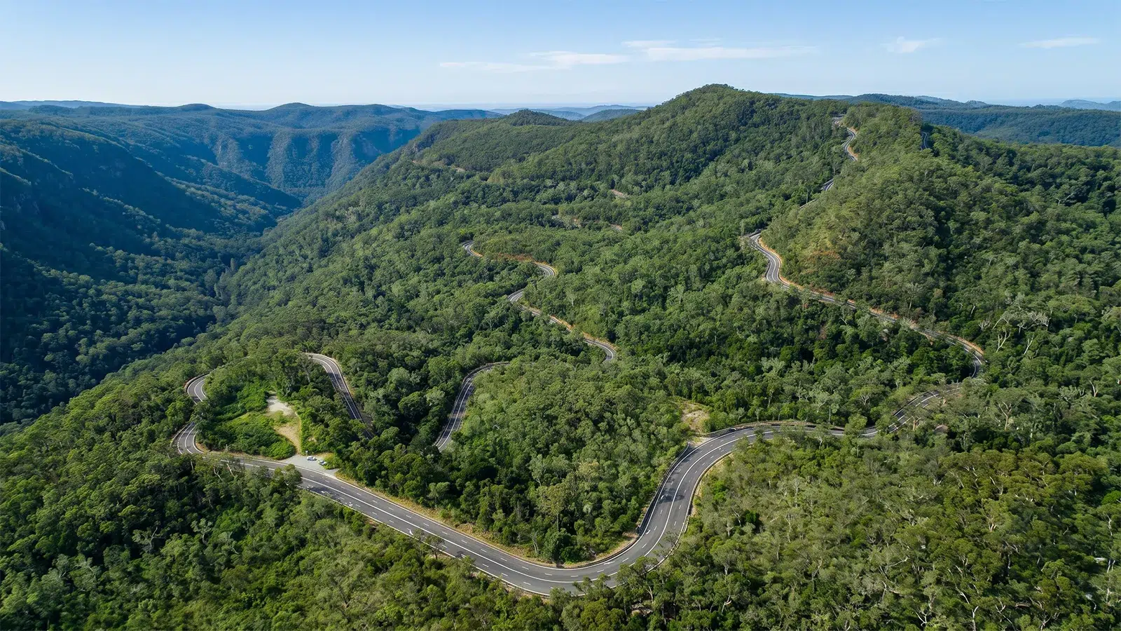 Aerial view of a sealed road switchbacking repeatedly through forested ranges under clear blue sky